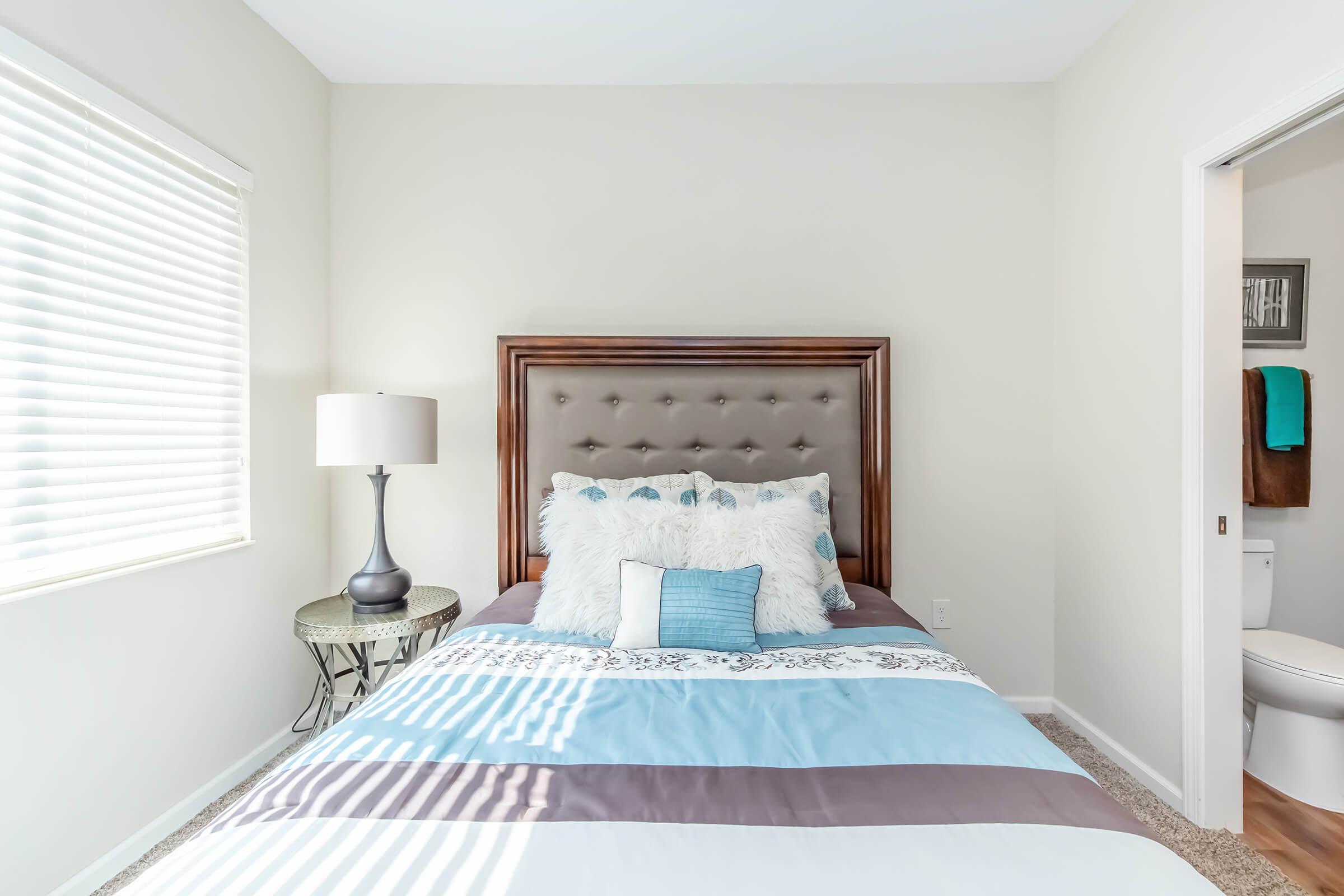 A cozy bedroom featuring a queen-sized bed with a tufted headboard, adorned with decorative pillows in shades of blue and white. A bedside table with a lamp sits next to the bed, and there is a window with blinds allowing natural light. A bathroom door is visible in the background.