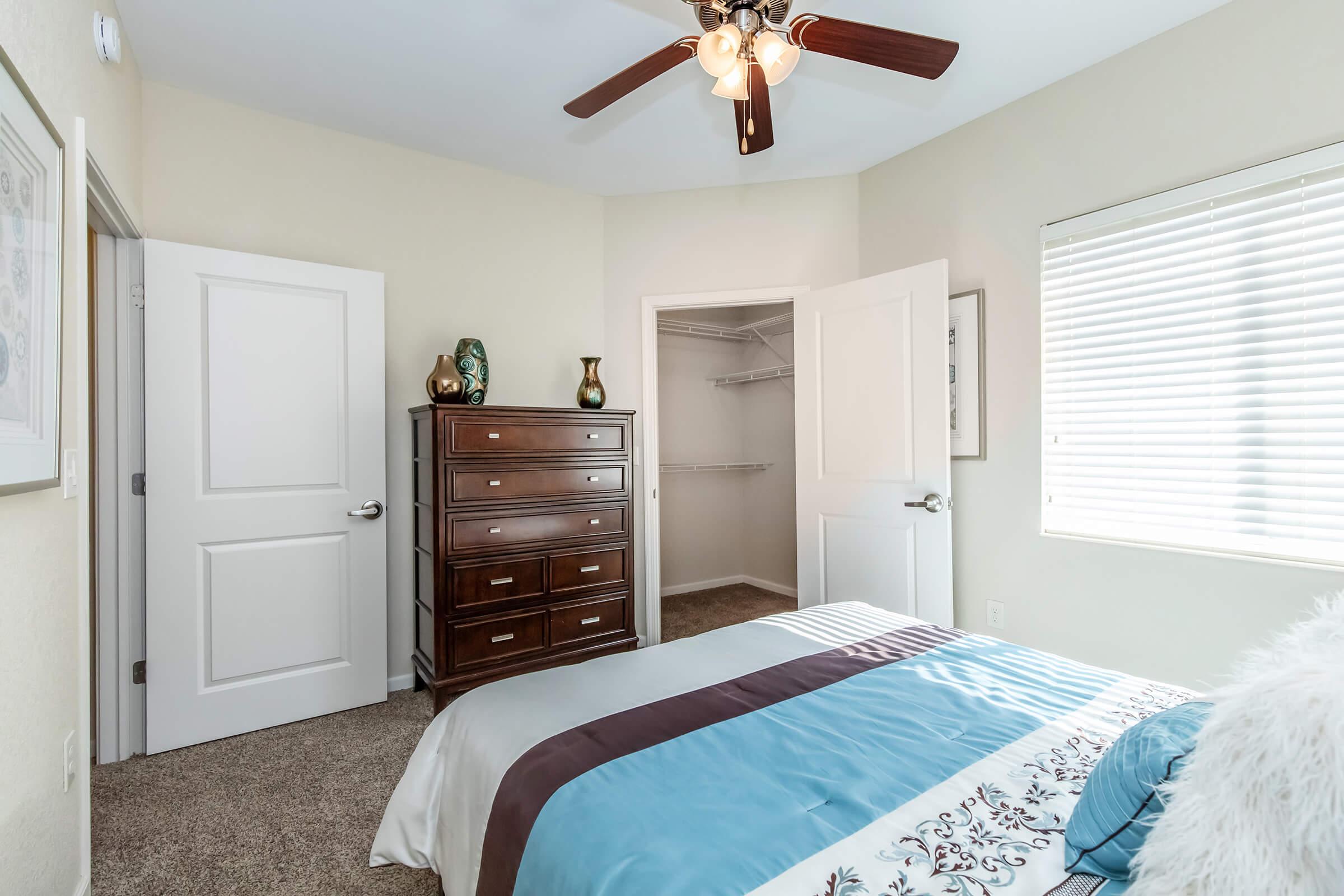 A cozy bedroom featuring a bed with a blue and white duvet, a wooden dresser with decorative items, and a walk-in closet. The room has light-colored walls, a ceiling fan with light fixtures, and a window with blinds allowing natural light to enter.