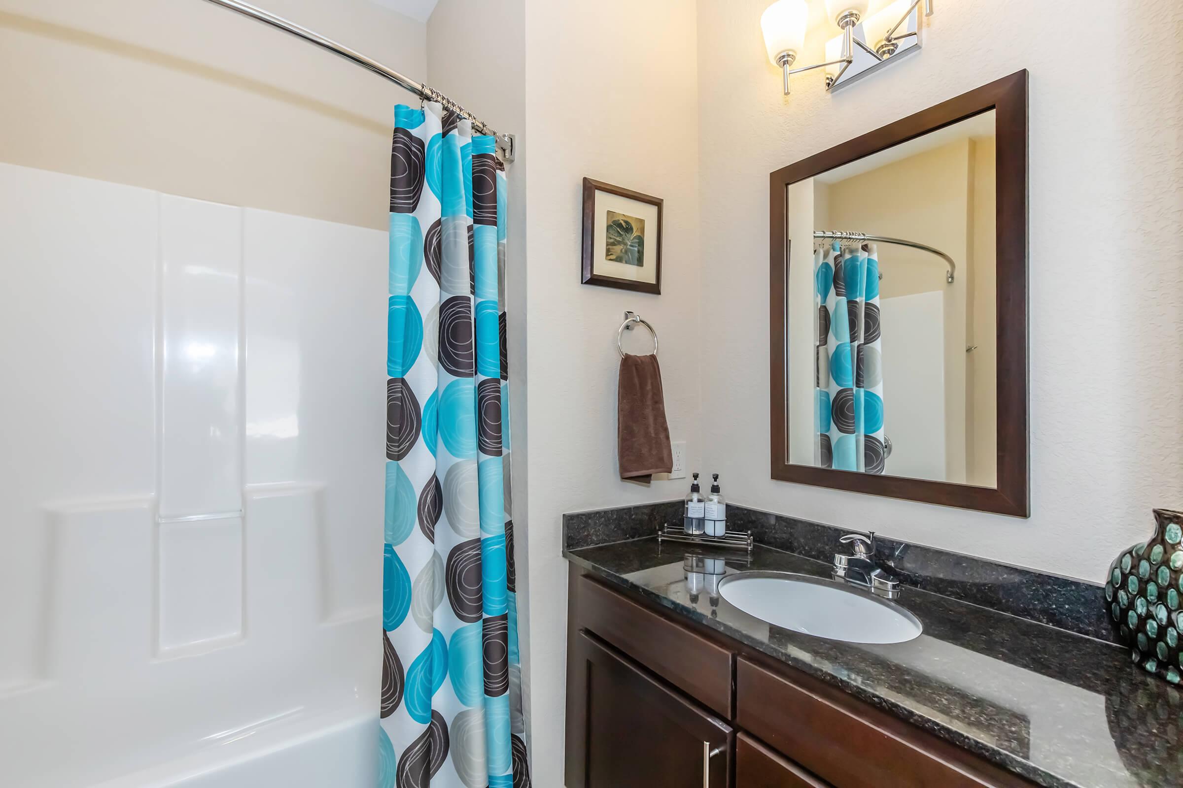 A modern bathroom featuring a shower with a white curtain, a dark wood vanity with a black countertop, and a circular mirror. Decorative elements include a blue and black patterned shower curtain, a towel on a rack, and toiletries arranged neatly by the sink. Soft lighting enhances the space.