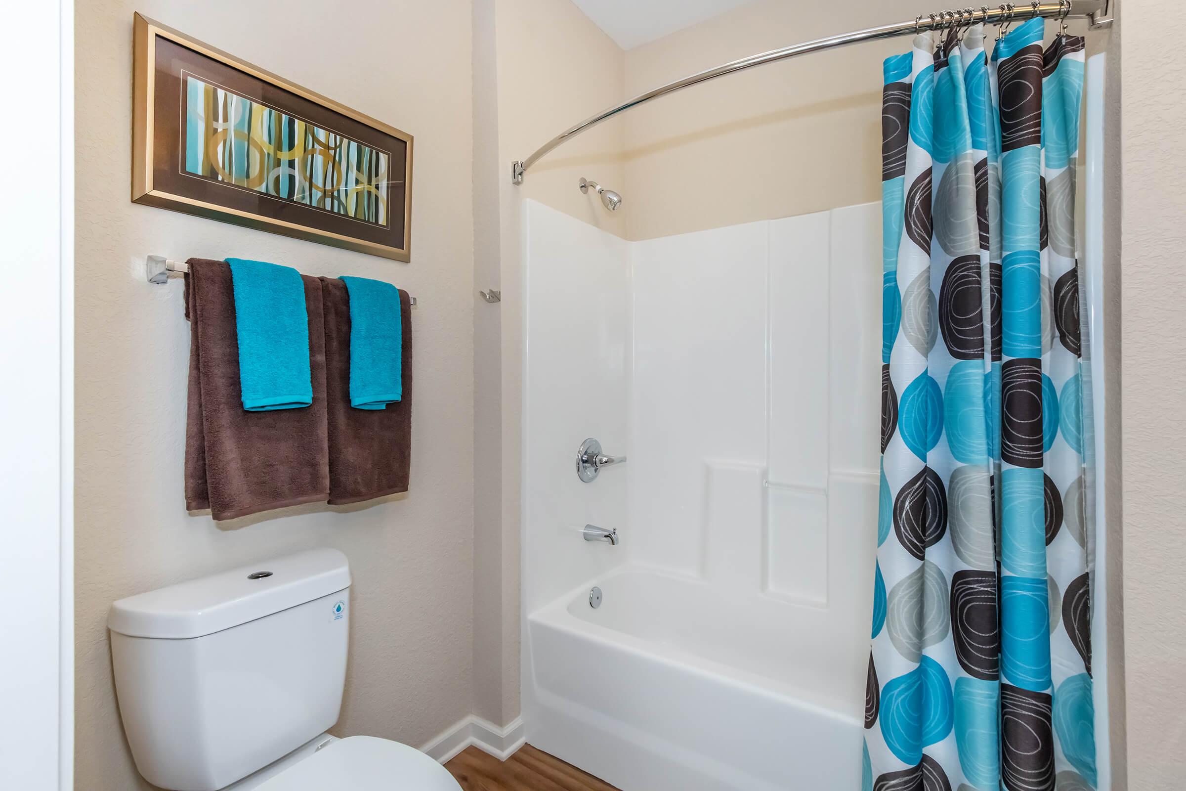 A modern bathroom featuring a white shower/tub combination, a decorative shower curtain with blue and brown circles, and two towels hanging on a towel rack: one teal and one brown. A clean white toilet is visible, along with light-colored walls and a framed artwork on the wall.
