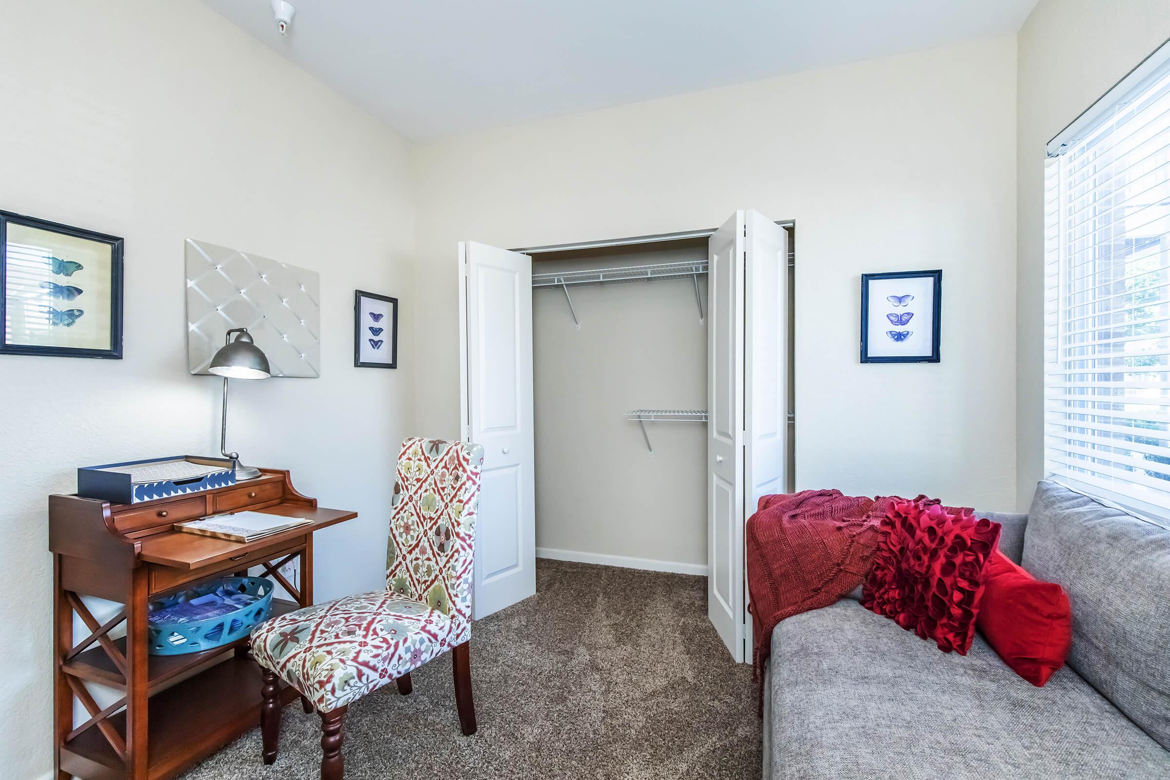 A cozy room featuring a small writing desk with a lamp, an upholstered chair, and a sofa with decorative pillows. Two open white doors lead to a closet with shelving. Walls are adorned with framed butterfly art, and the space has light-colored walls and carpeting, creating a bright, inviting atmosphere.