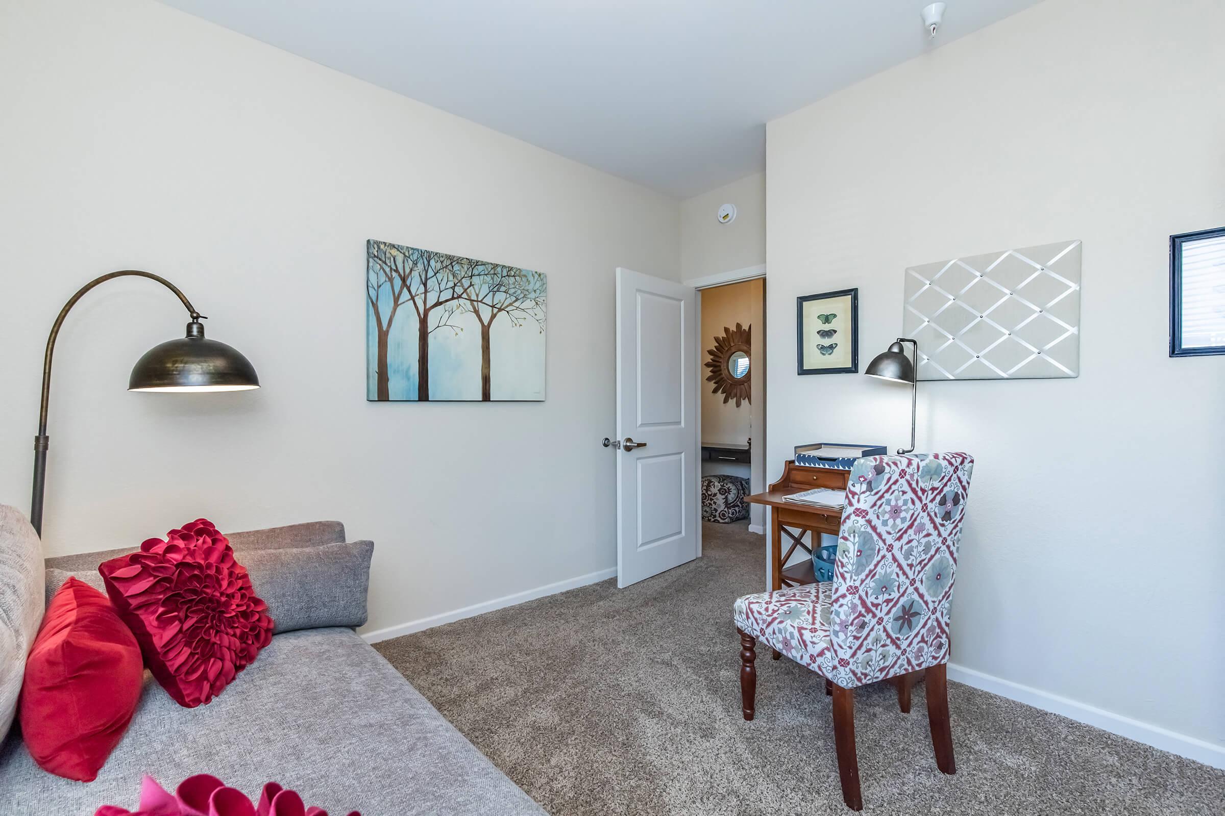 A cozy room featuring a gray sofa with red accent pillows, a stylish floor lamp, and a patterned chair. Two framed artworks depicting trees and a mirror adorn the light-colored walls. A door leads to another space, and a small desk can be seen in the background. The carpet is plush and soft.