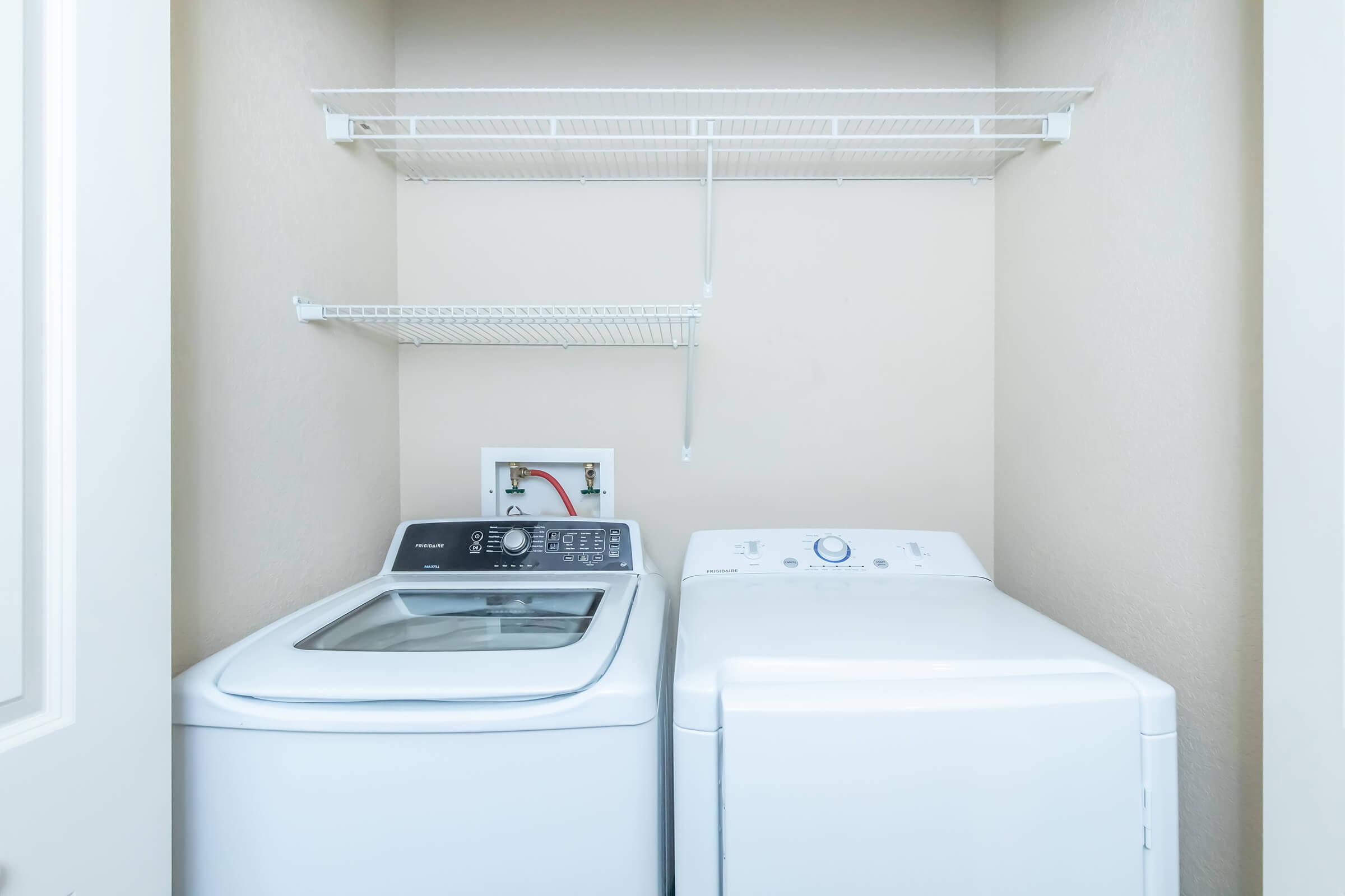 A small laundry room featuring a top-loading washing machine on the left and a front-loading dryer on the right. Above the appliances, there is a wire shelving unit against a neutral-colored wall, providing storage space. The floor is clean and unobstructed.