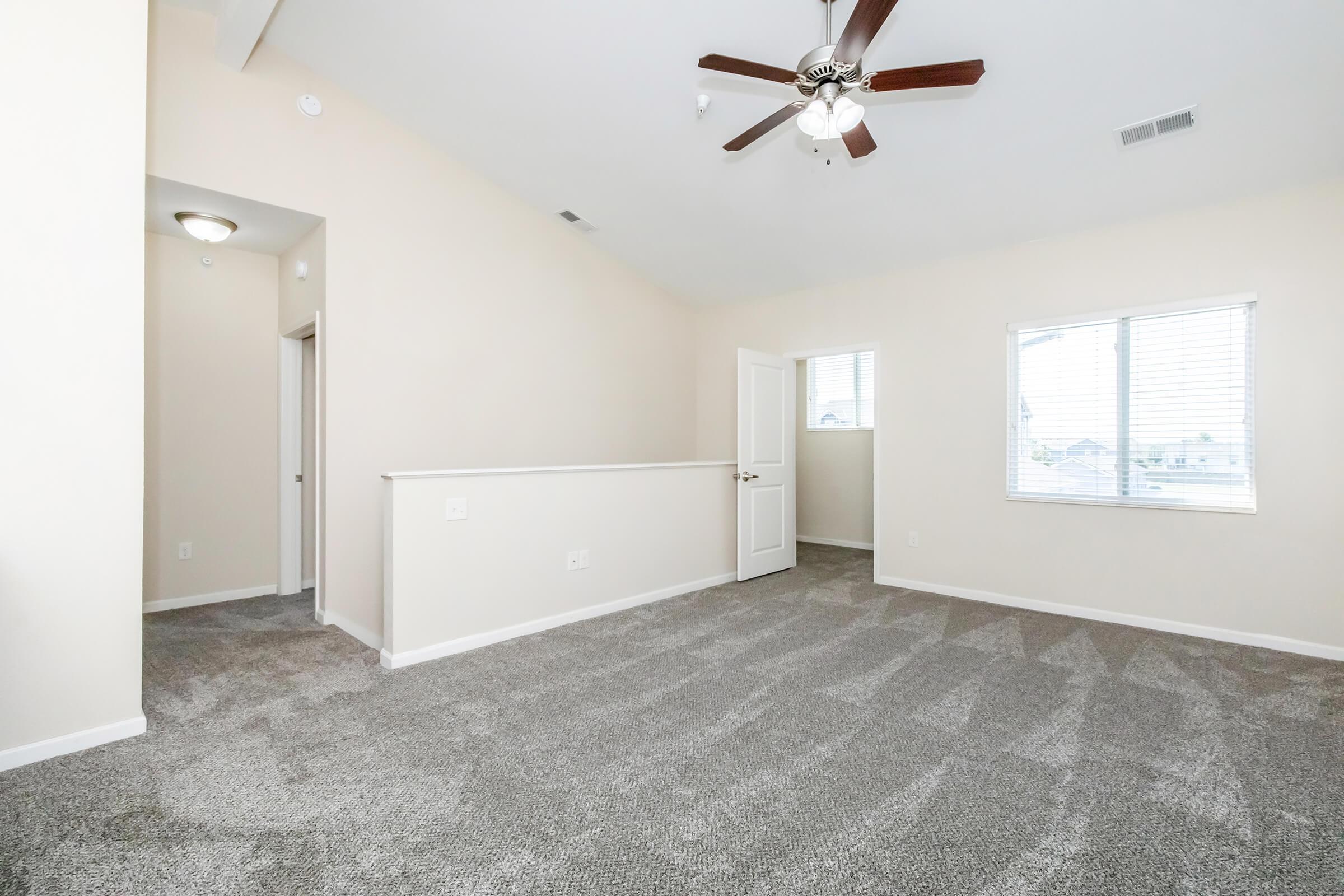 Spacious room with light-colored walls and carpeted flooring. A ceiling fan hangs from the high ceiling, and there are two windows allowing natural light. A door on the left leads to another space, and the room has a bright, airy feel.