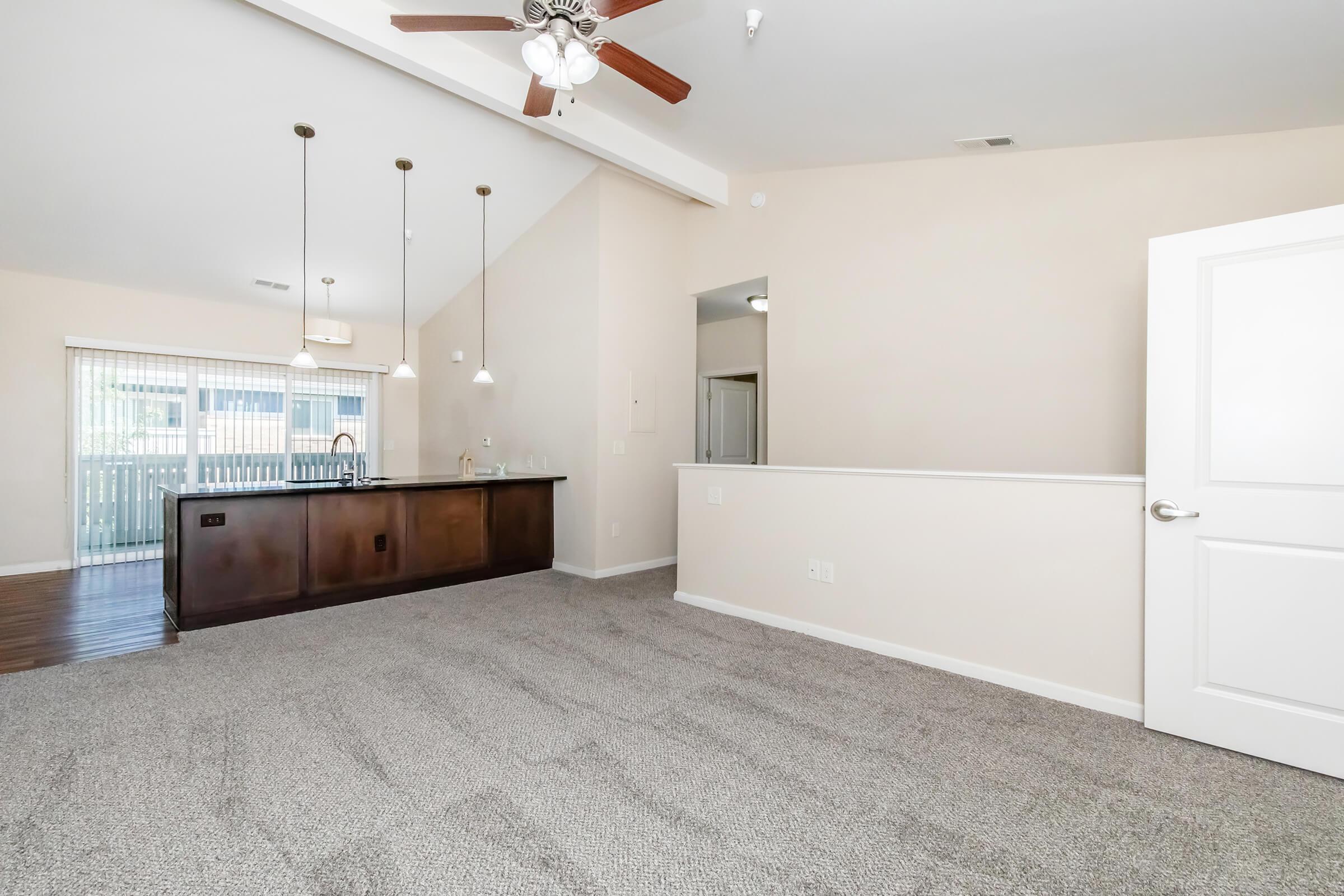 Spacious living area with a ceiling fan, featuring light-colored walls and carpeted flooring. A kitchen bar with pendant lighting is in the background, leading to a dining space with sliding doors. Natural light enters from the windows, creating a welcoming atmosphere.