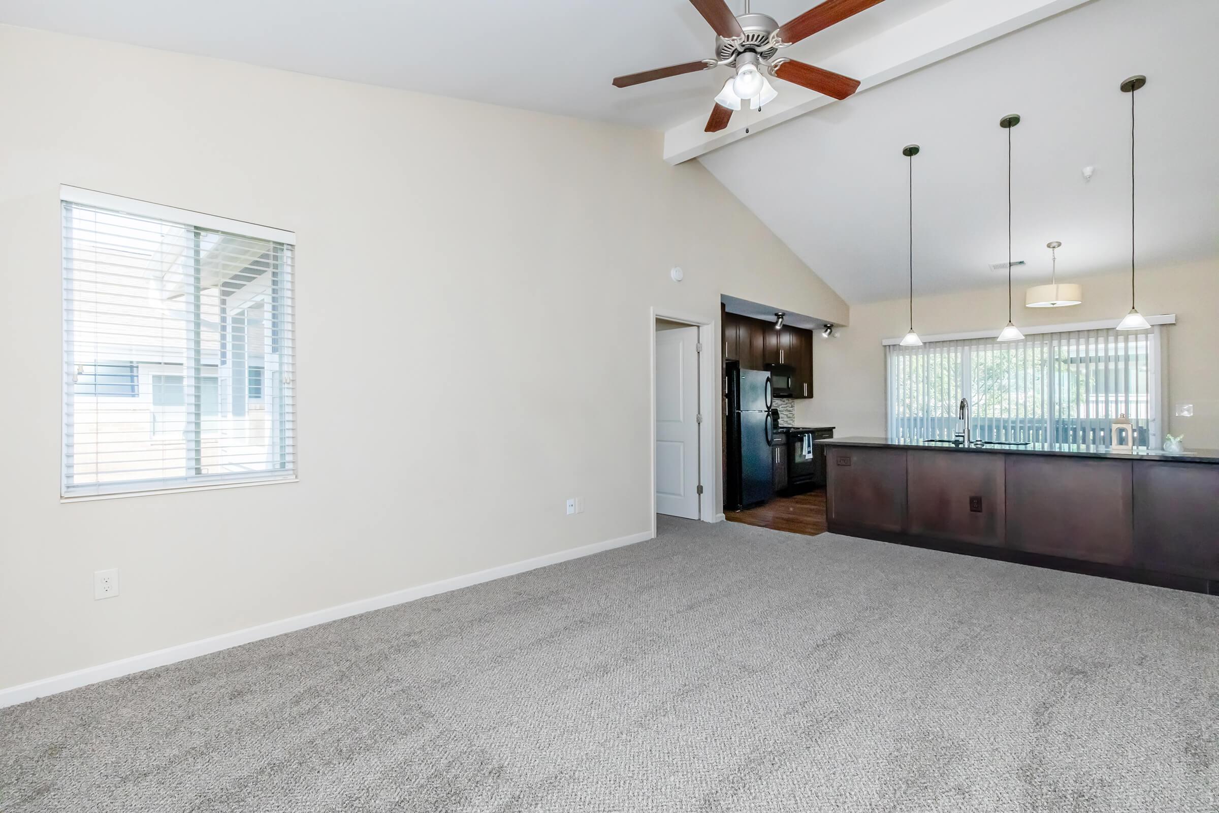Spacious living area with carpeted flooring, featuring a ceiling fan and natural light from a window. A doorway leads to a kitchen area with dark cabinetry and pendant lighting, and a large countertop. The walls are painted in light tones, creating a bright and inviting atmosphere.