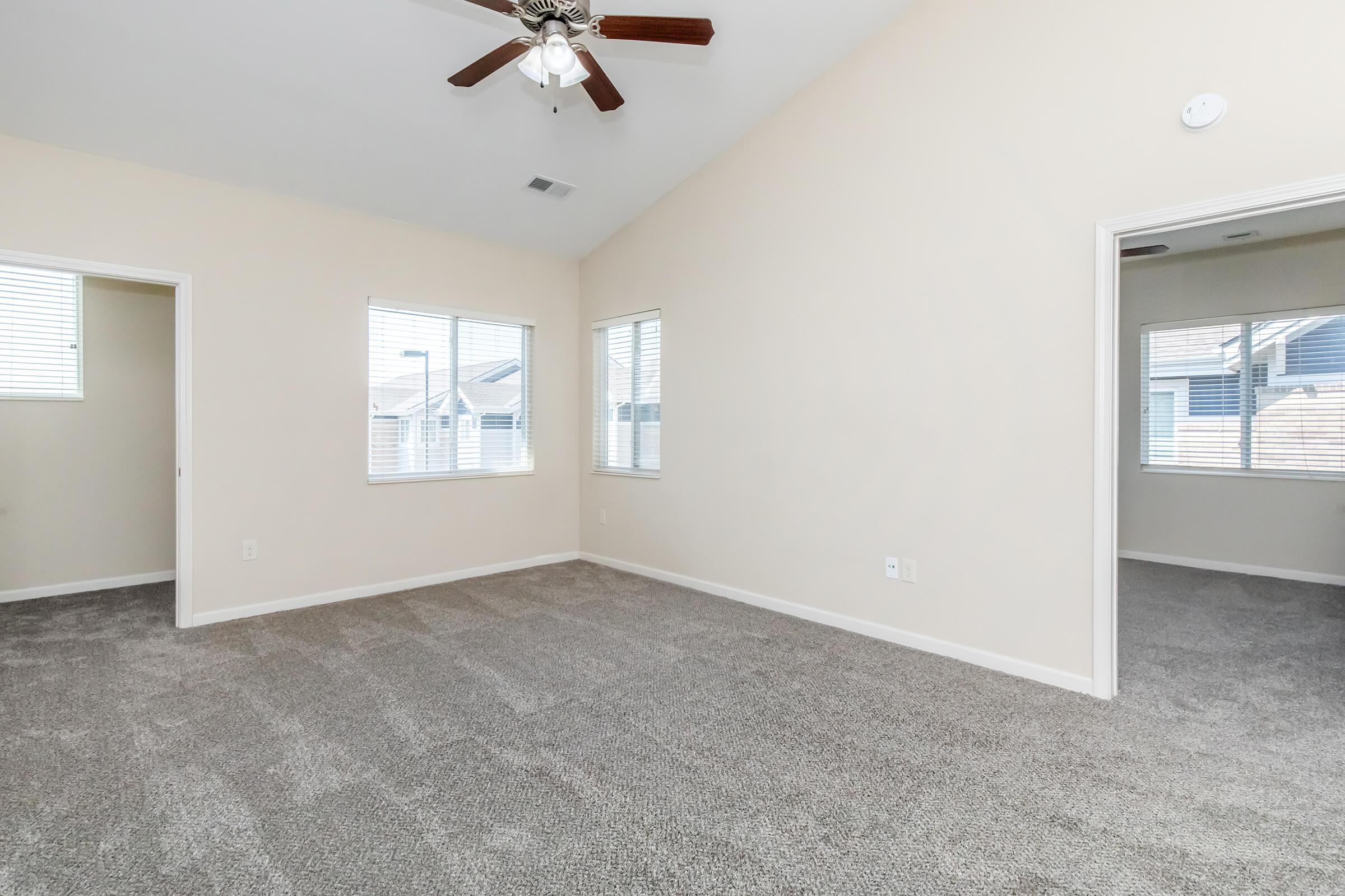 Spacious room with freshly carpeted flooring, large windows allowing natural light, and a ceiling fan. Two doors lead to adjacent rooms, creating a bright and airy atmosphere. Neutral wall color enhances the openness of the space.