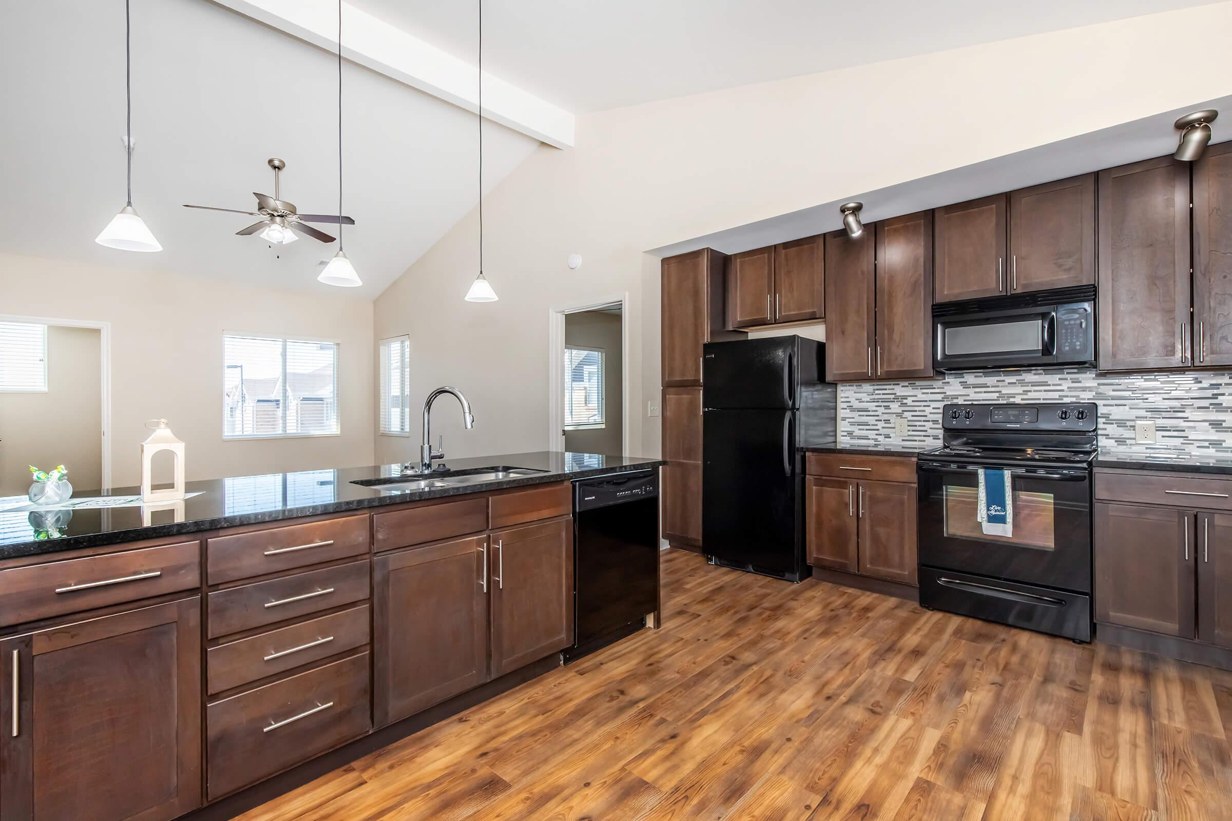Modern kitchen featuring dark wood cabinets, a black countertop, and stainless steel appliances including a stove, microwave, and refrigerator. The space has recessed lighting, a ceiling fan, and large windows allowing natural light to fill the room. The floor is made of light-colored wood.
