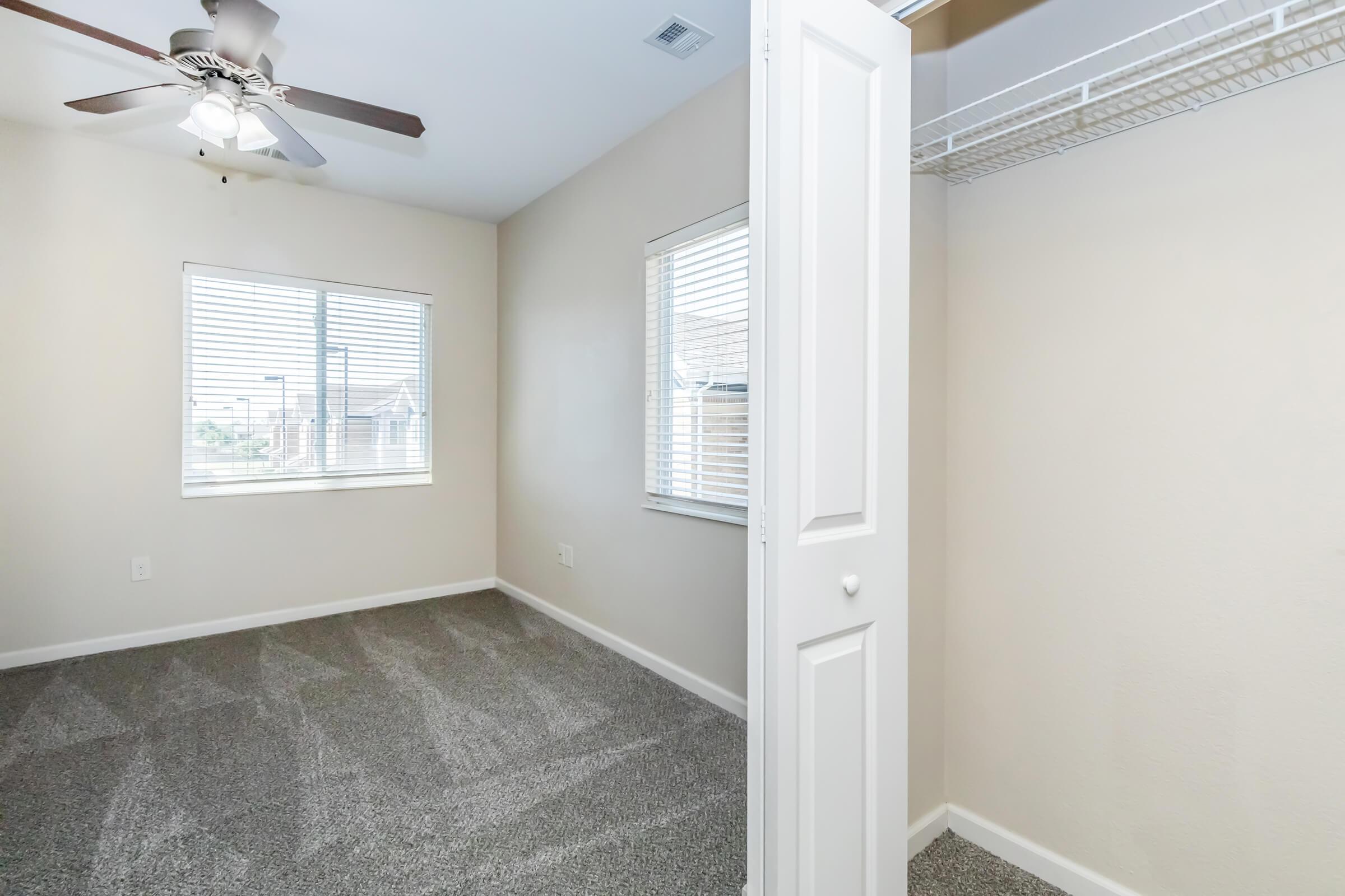 A well-lit room featuring a ceiling fan and two windows with blinds. The room has light-colored walls and is carpeted in a soft gray. In the foreground, there is an open closet with a white door, showcasing a wire shelf. The overall ambiance is clean and bright, suitable for personal or office space.