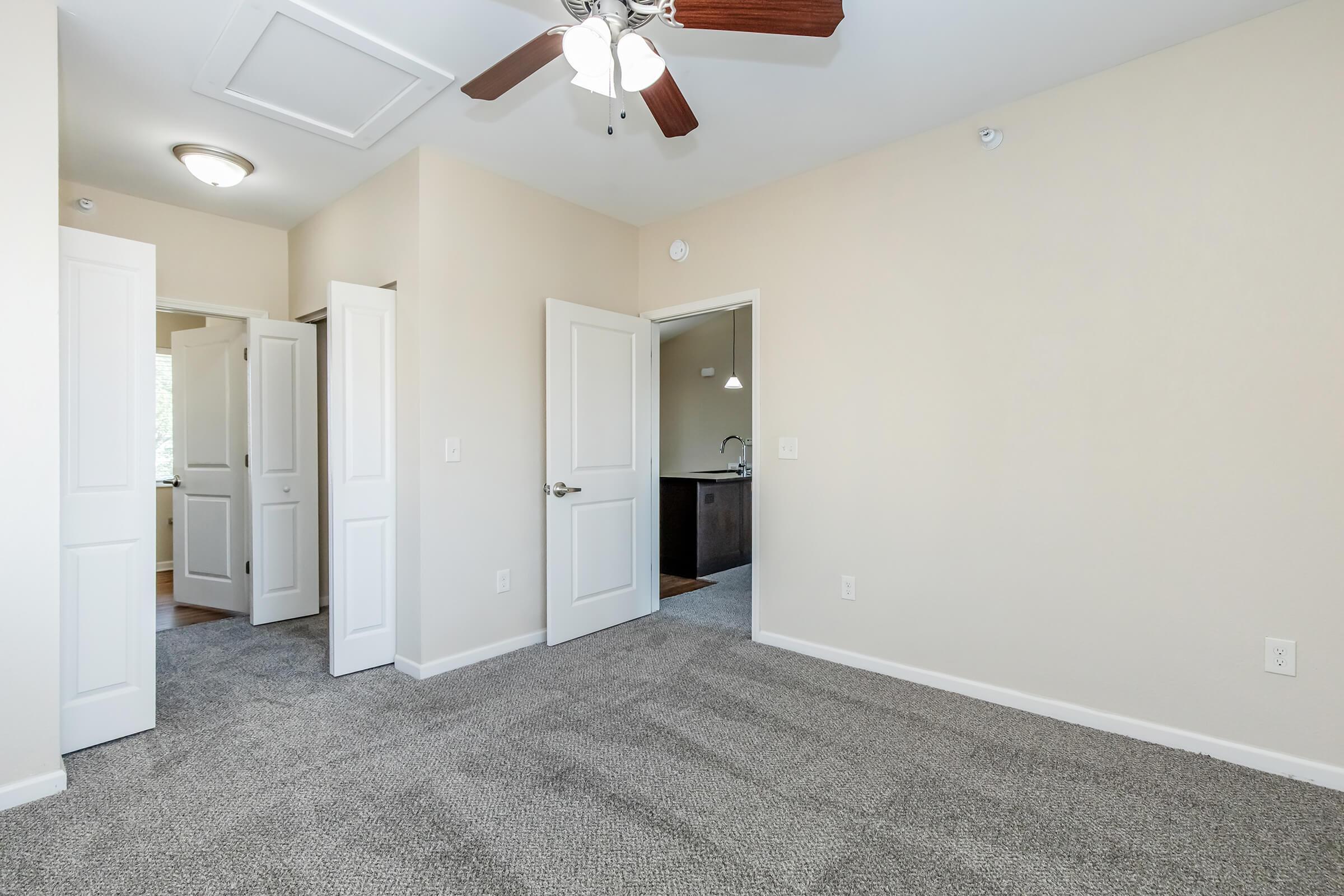 A spacious room with light beige walls and a ceiling fan. It features soft gray carpet, two white doors leading to adjacent areas, and natural light coming from a window partially visible in the background, showcasing a modern kitchen area.