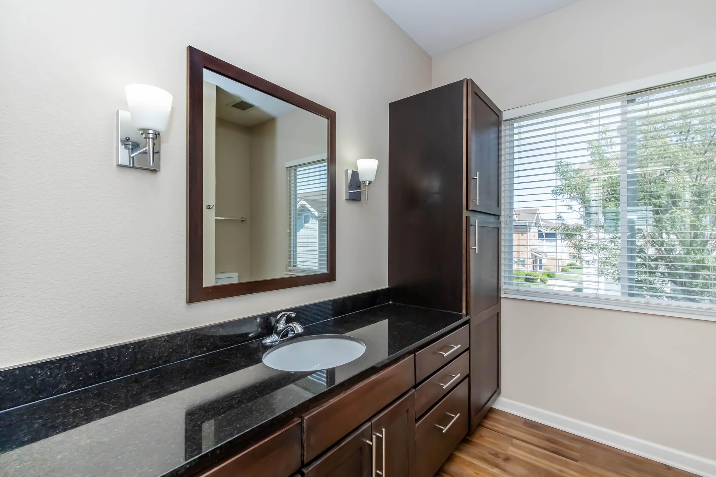 A modern bathroom featuring a dark wood vanity with a black countertop and a circular sink. A large mirror is mounted above the sink, and there are two stylish wall sconces on either side. A window with white blinds lets in natural light, and the flooring is hardwood.