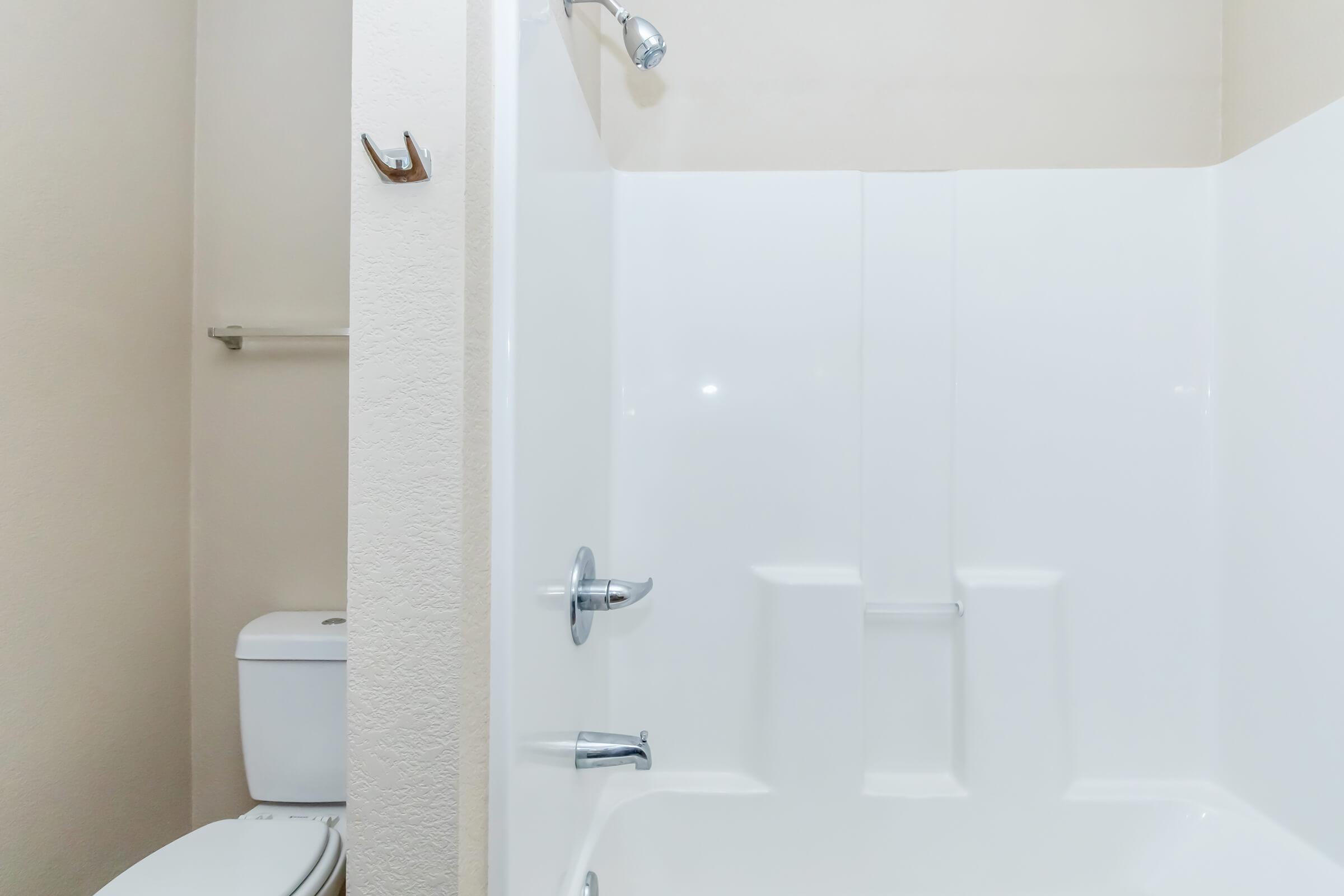 A clean, well-lit bathroom featuring a white bathtub with a showerhead, and a white toilet nearby. The walls are a light, neutral color, and there’s a small shelf attached to the wall, enhancing the simple, modern design of the space.