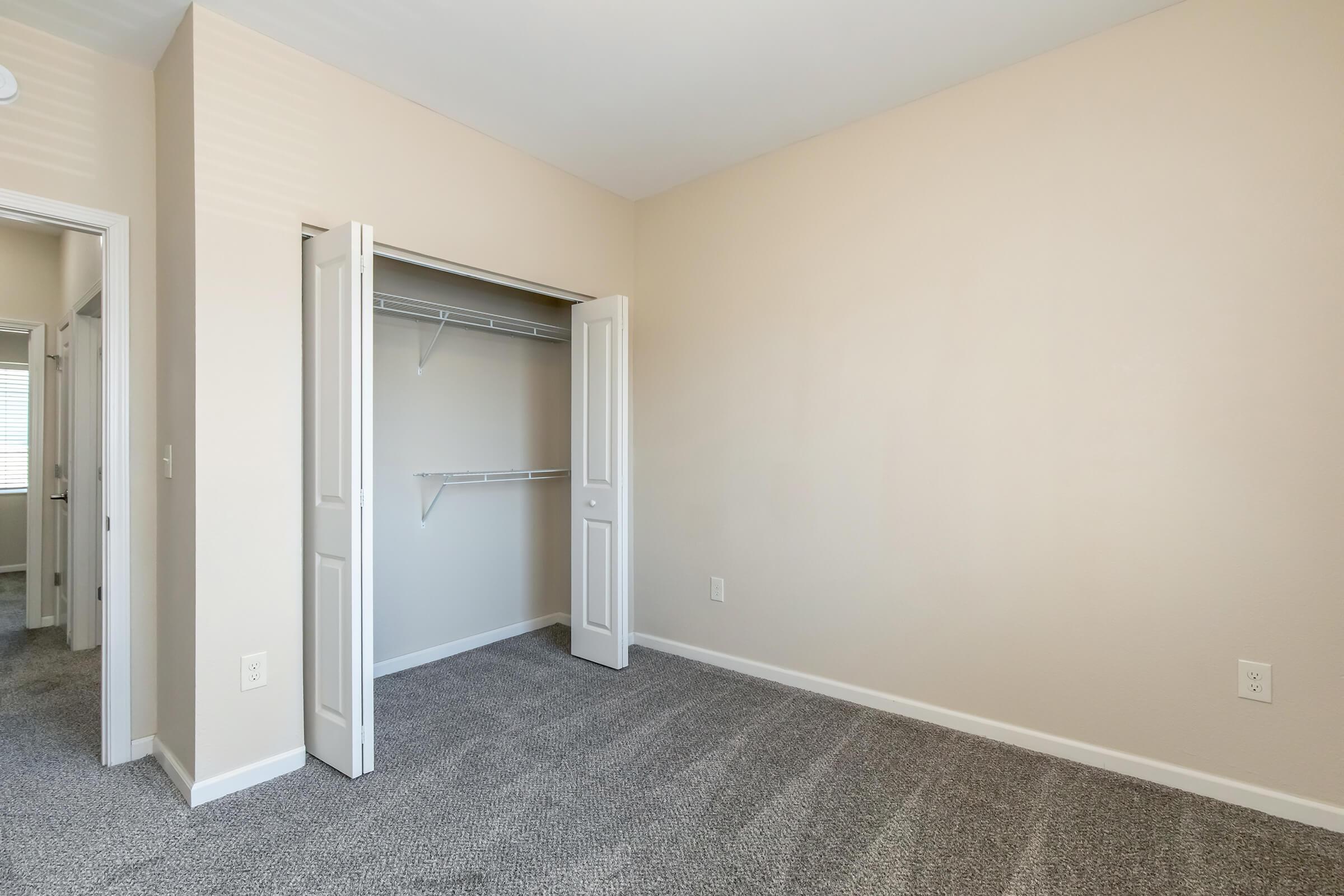 A vacant room with light beige walls and gray carpet. There is an open closet with shelving on the left side, and two doorways leading to other areas. The room is well-lit and spacious, creating a neutral and inviting atmosphere.