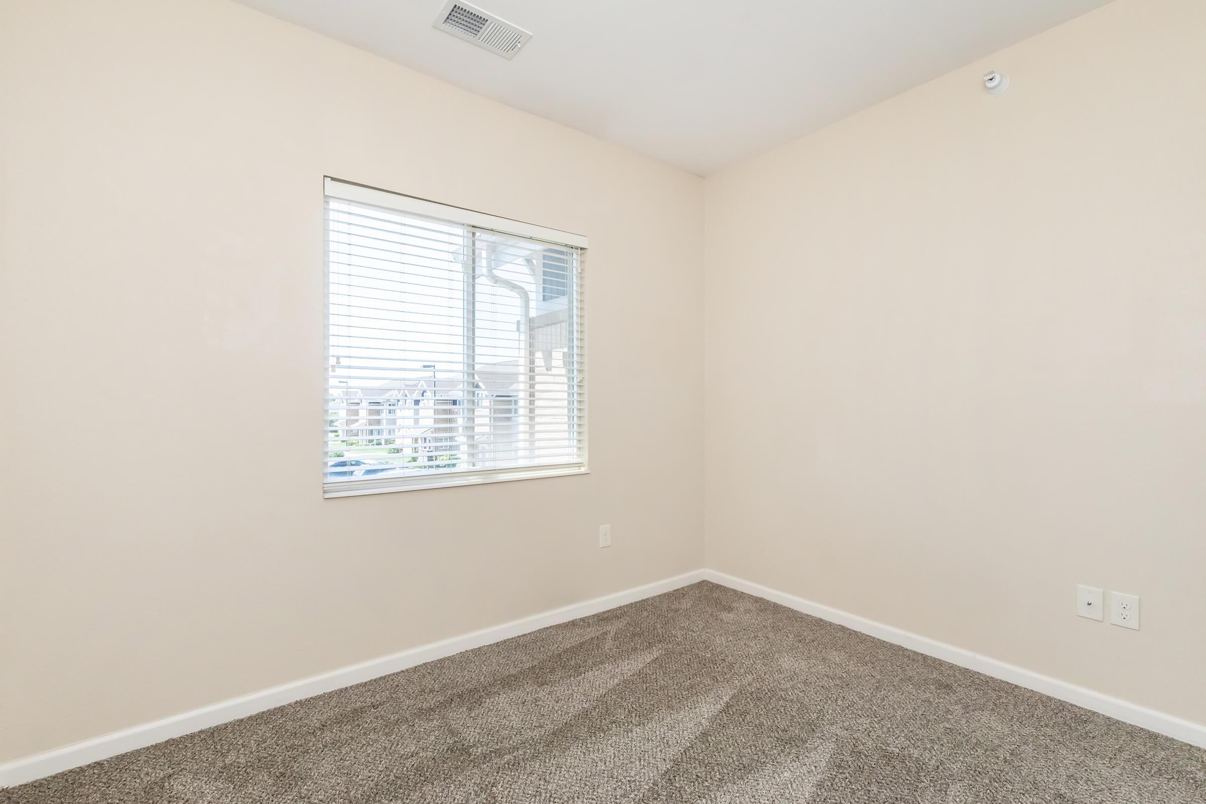 Empty room with beige walls and light-colored carpet. A window with white blinds lets in natural light, revealing a view of buildings outside. The room features minimal decor and is clean and tidy, with an electrical outlet on one wall.