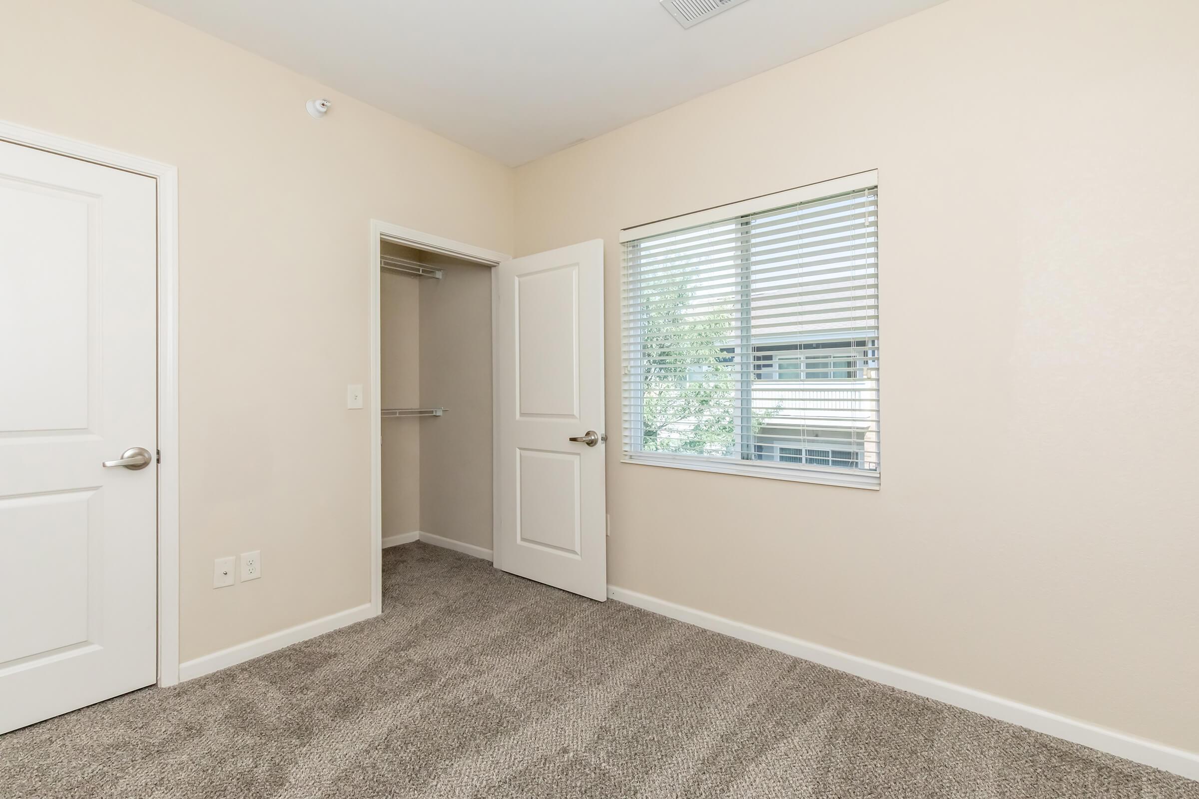 A light-colored room with beige walls and carpet. It features a closed white door leading to a closet on the left, another white door to the right, and a window with white blinds allowing natural light to enter the space.