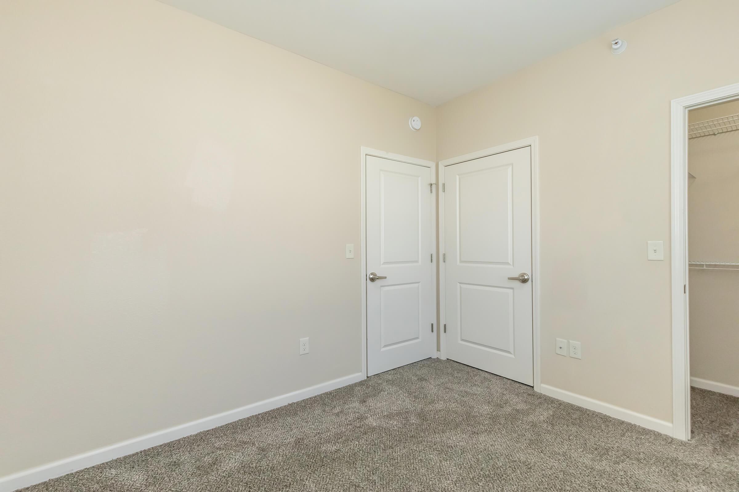 A well-lit room with beige walls and light gray carpet. There are two white doors on the right side, one leading to a closet. The space is empty, creating a clean and minimalist appearance, suitable for various uses such as a bedroom or office.
