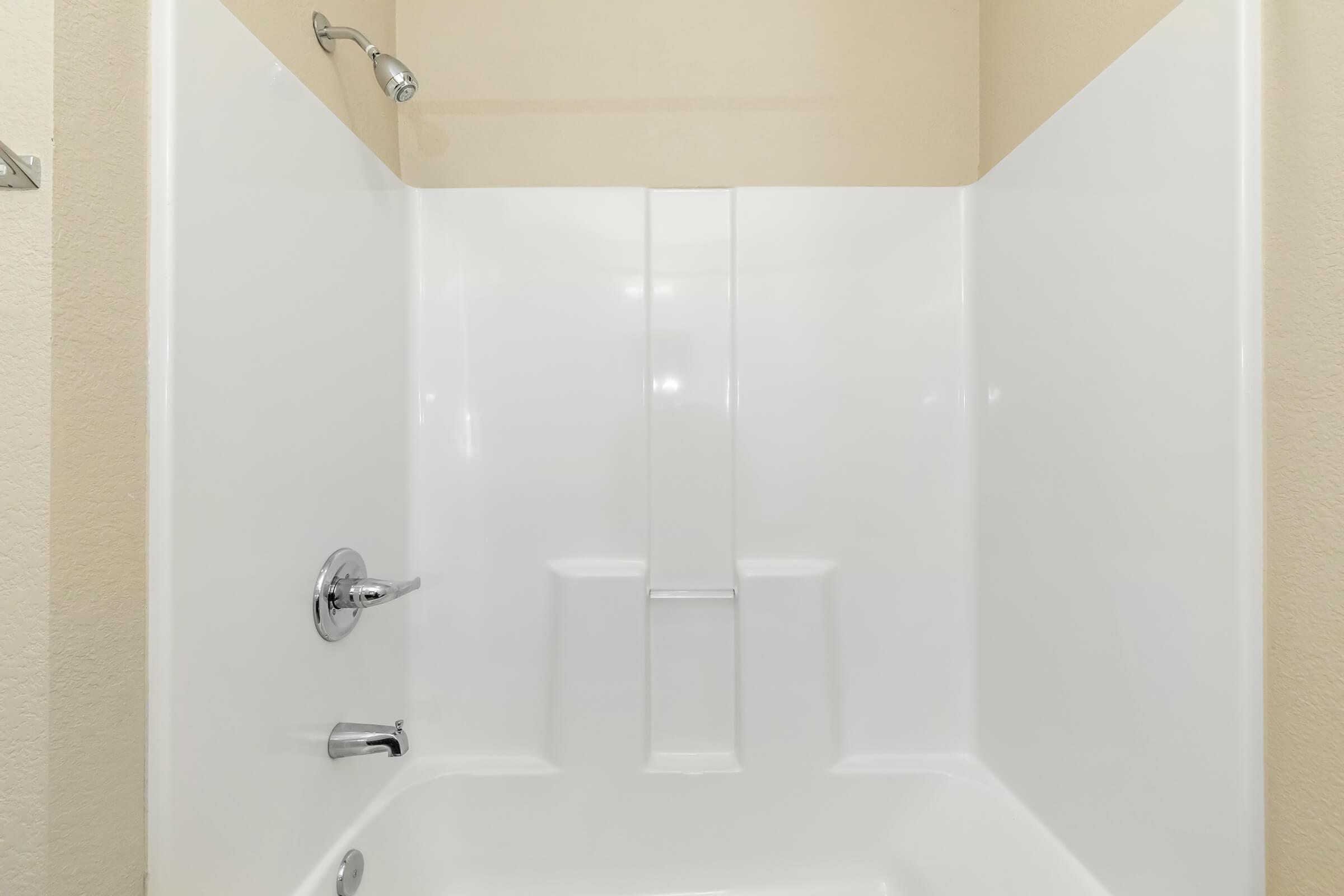 A clean, minimalist shower stall with a white acrylic surround and a chrome showerhead. The design features a built-in shelf and a smooth, seamless appearance, set against a light beige wall, creating a bright and spacious feel.
