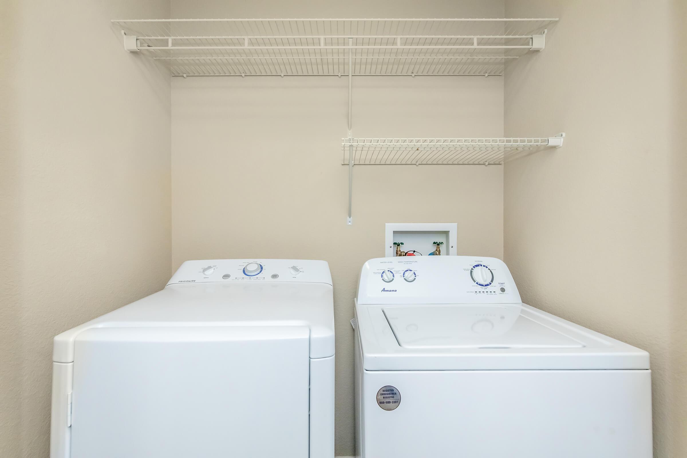 A laundry room with two white appliances: a washing machine on the left and a dryer on the right. Above them, there is a wire shelf for storage. The walls are painted a neutral color, creating a clean and organized space.