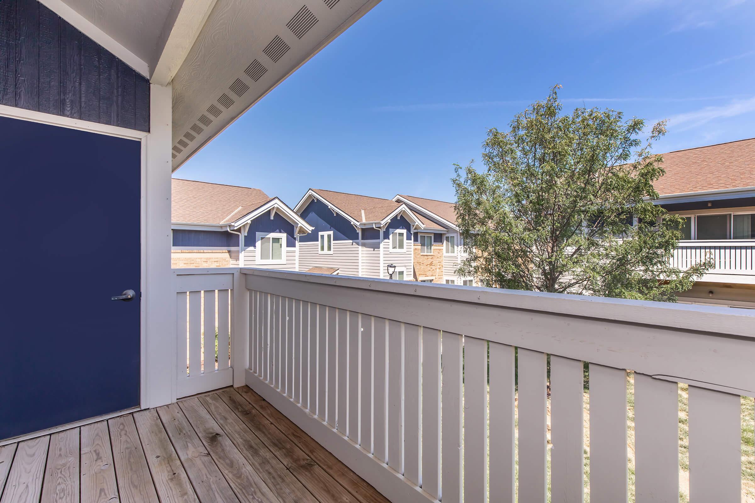 A view from a balcony featuring a wooden deck, white railing, and a blue door. In the background, there are buildings with pitched roofs and a tree. The sky is clear with a few clouds, creating a bright, sunny atmosphere.