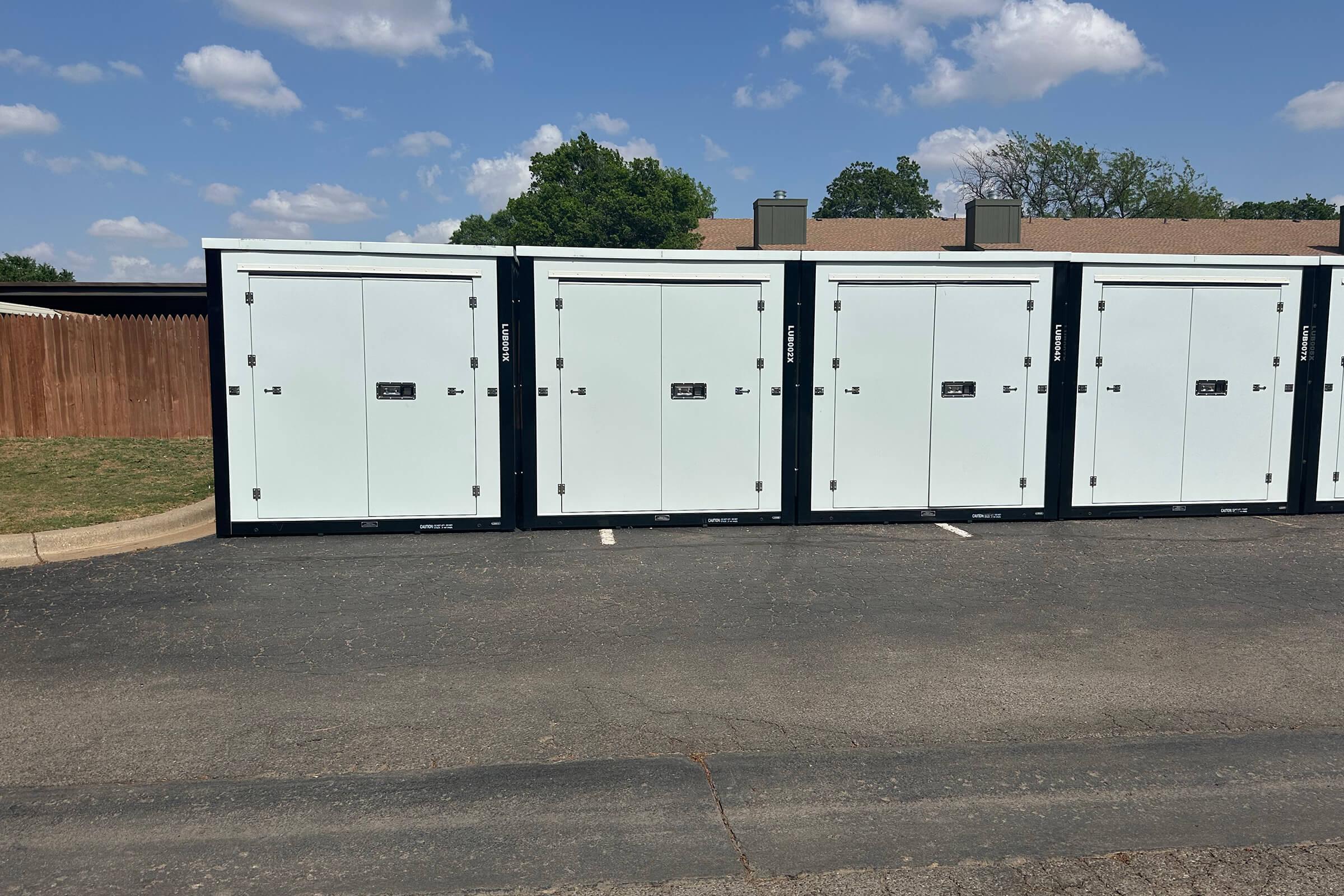 Row of storage units featuring white doors and black frames, lined up on a paved surface. A wooden fence is visible in the background, with blue skies and clouds above. The setting appears to be a storage facility.