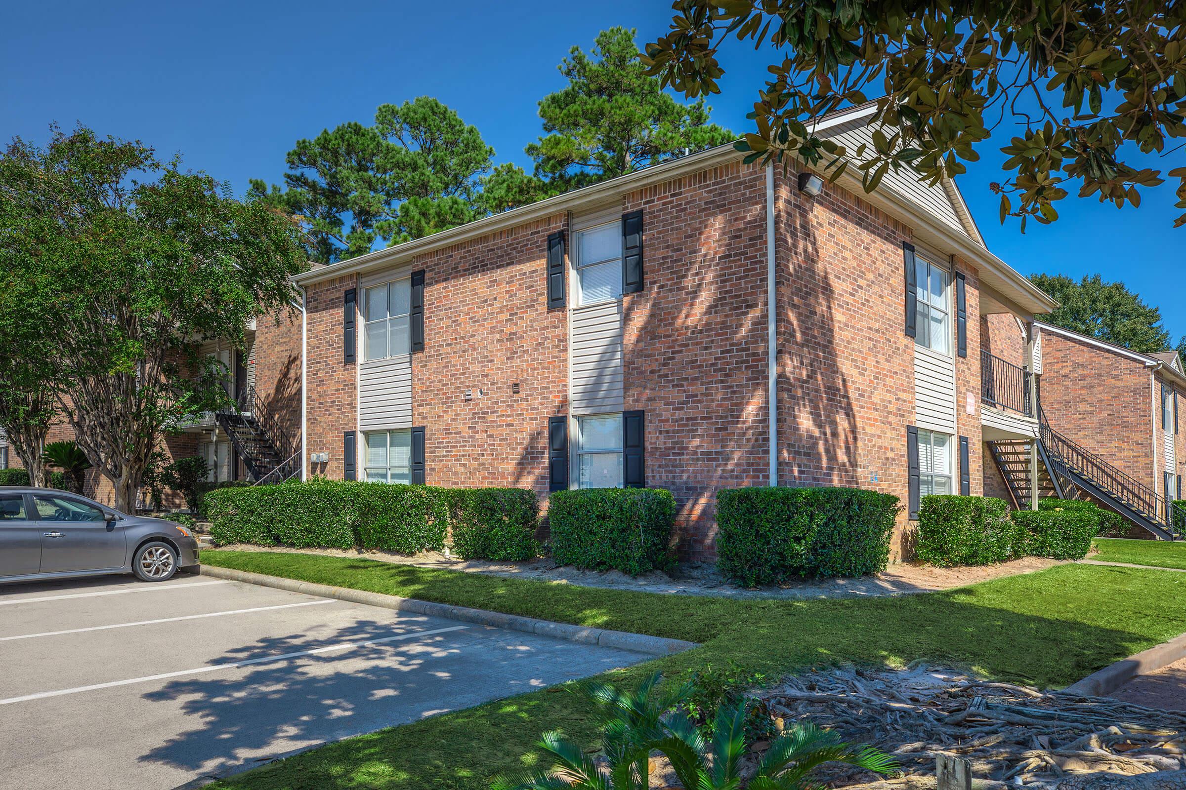 A two-story brick apartment building surrounded by green shrubs and trees, with a clear blue sky above. In the foreground, there is a parking lot with a single parked car. The building features several windows and stairs leading to upper units.