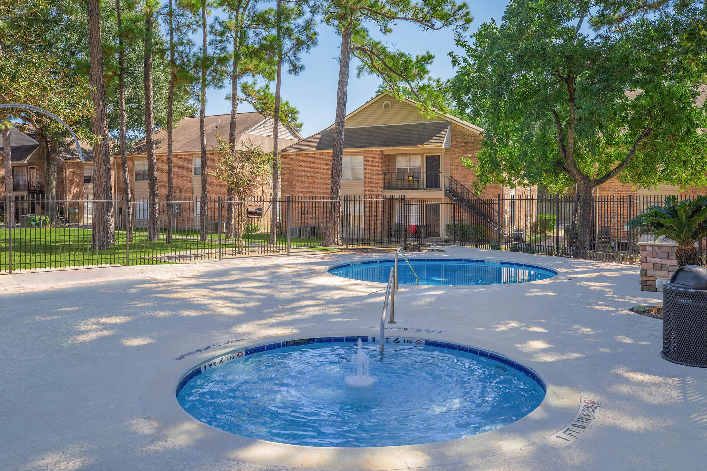 A serene outdoor area featuring two circular hot tubs surrounded by a patio. Lush green trees provide shade, and there are apartment buildings in the background. The space is enclosed with a fence, creating a peaceful atmosphere for relaxation.