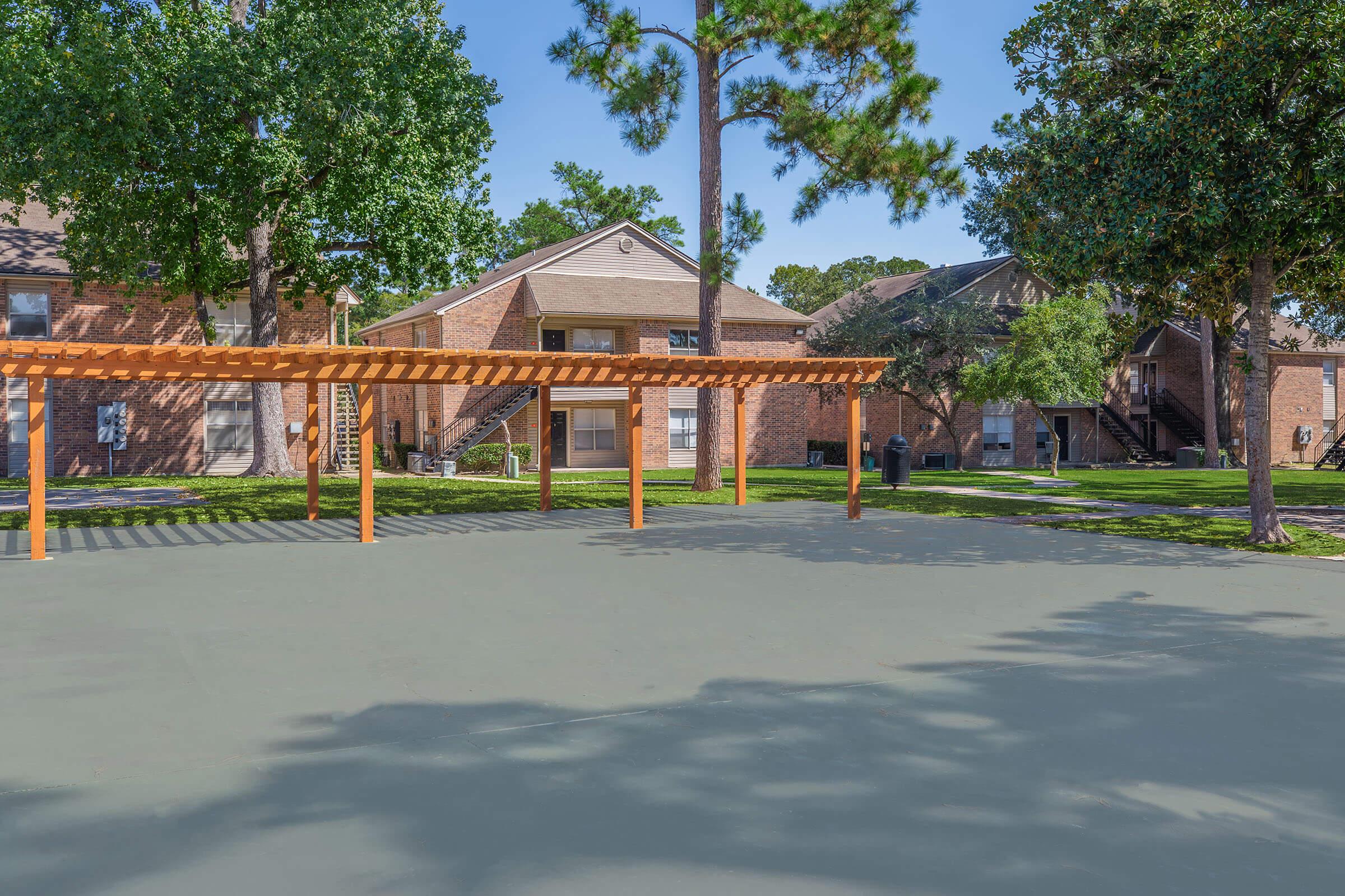 A sunny outdoor area featuring a shaded pergola with wooden beams, surrounded by grass and trees. Nearby, there are brick apartments and a clear blue sky. The scene is tranquil and inviting, perfect for relaxation or social gatherings.