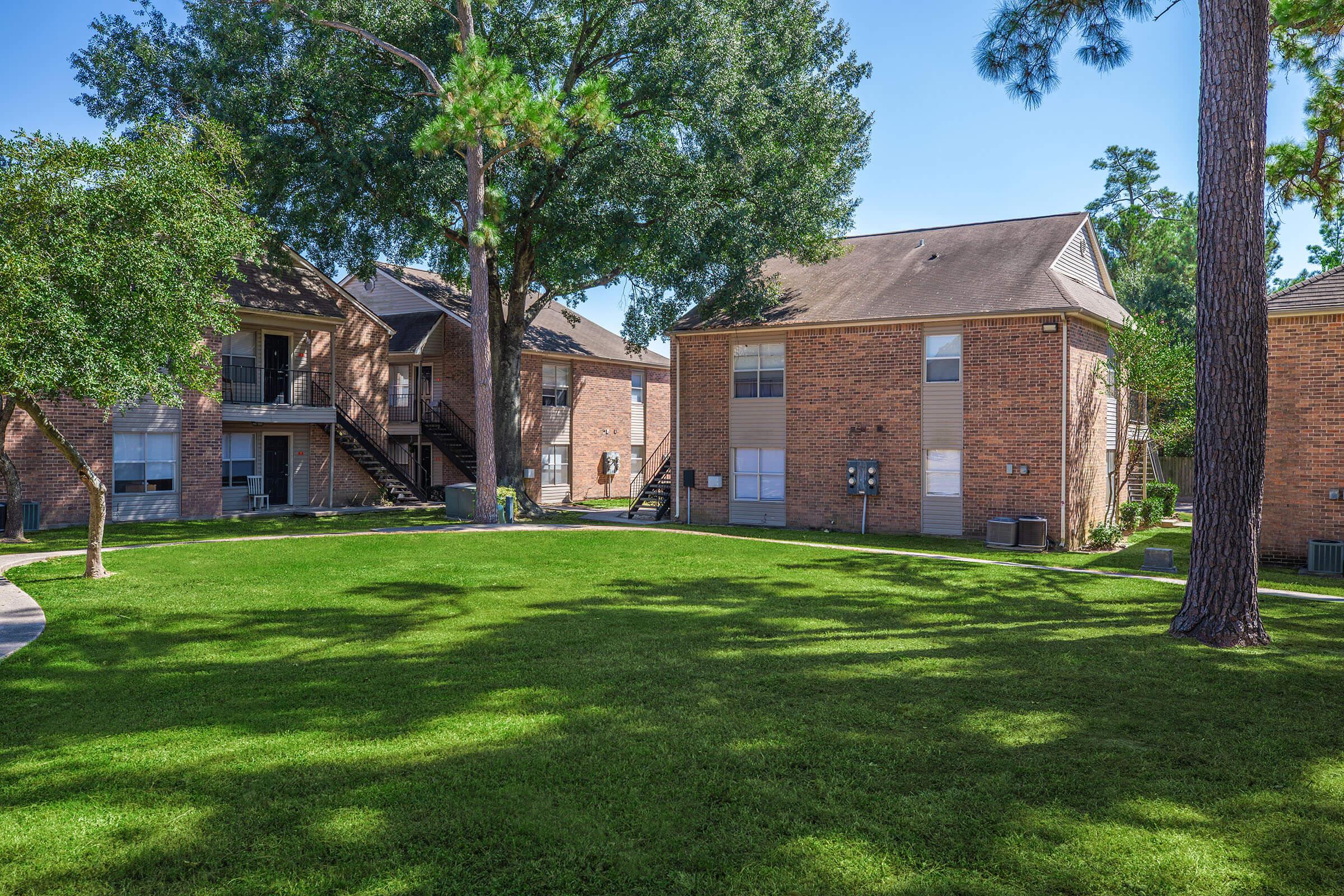 A landscaped apartment complex featuring two brick buildings surrounded by lush green grass and trees. The scene includes clear blue skies and well-maintained walkways leading to the entrances.