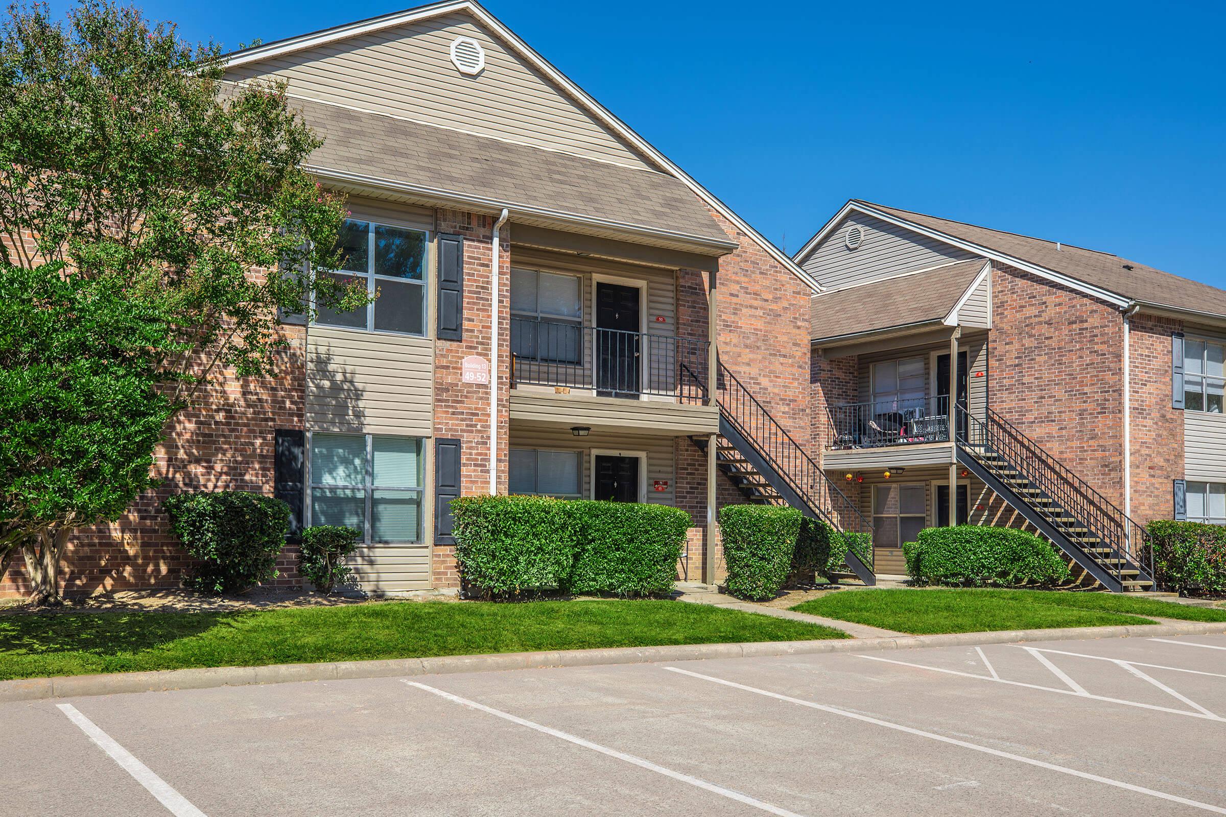 Multifamily residential buildings featuring tan and brick exteriors, two stories with balconies, surrounded by manicured greenery. The parking lot in front shows marked spaces, and a blue sky in the background enhances the setting.