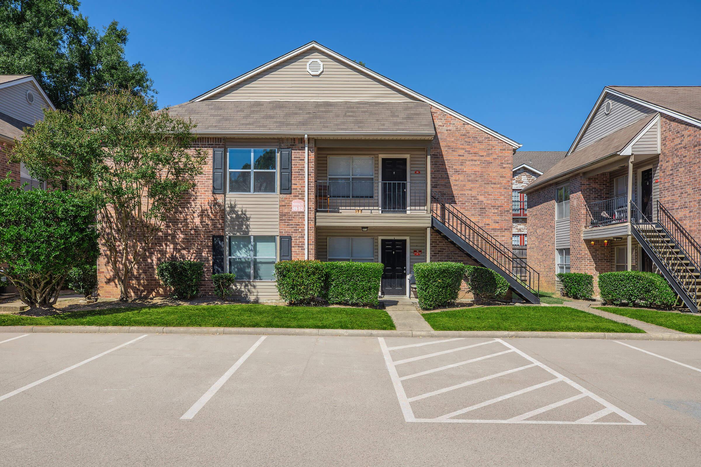 Two-story brick apartment building with a landscaped entrance. The facade features large windows and balconies. There are two sets of stairs leading to the second floor. In front, a well-maintained parking lot with marked spaces and green shrubs lining the pathway. Clear blue sky overhead.