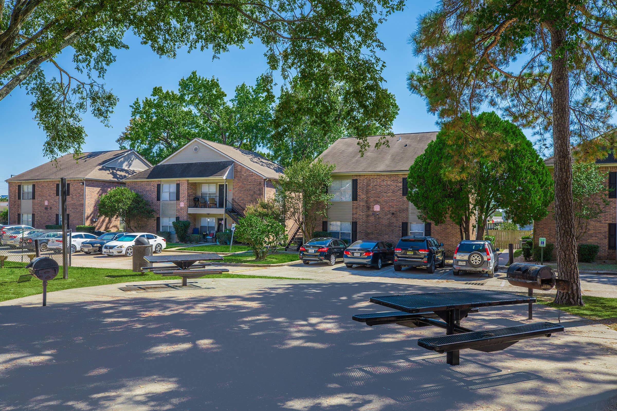 A residential complex with brick buildings surrounded by trees. In the foreground, there are picnic tables and charcoal grills. Several cars are parked nearby, and the area appears well-maintained and inviting, with clear blue skies overhead.