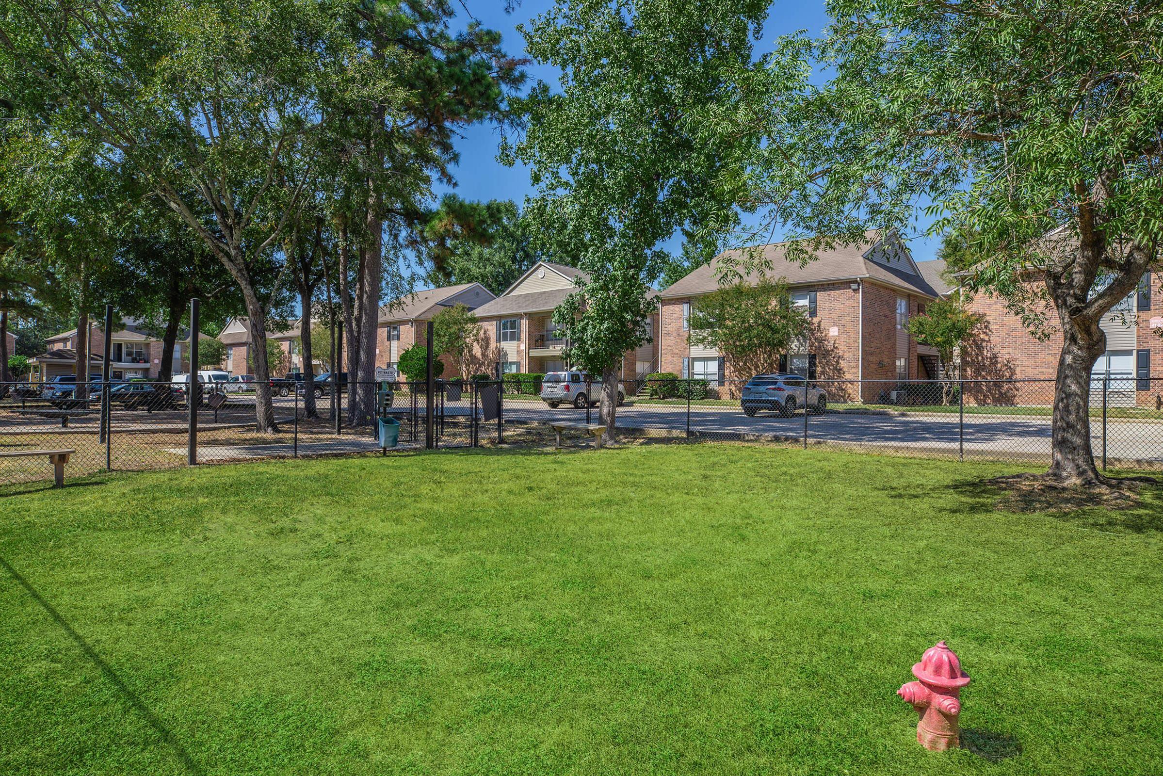 A well-maintained green lawn in a park-like setting, with a bright pink fire hydrant in the foreground. Behind the lawn are several residential buildings, surrounded by trees and a black fence. Vehicles are parked nearby, indicating a residential area.
