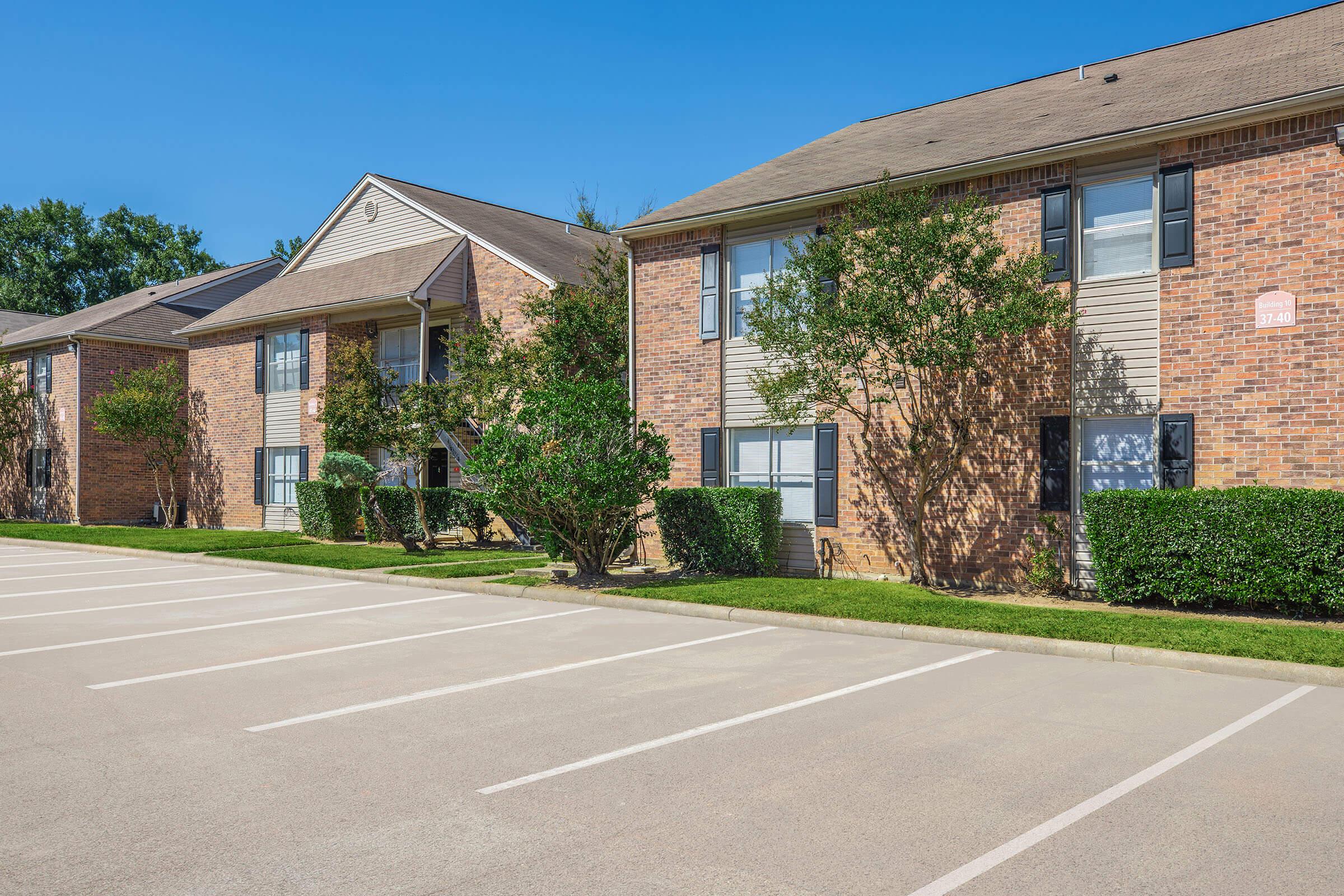 A row of multi-unit brick apartment buildings with pitched roofs and trees in front. The parking lot in the foreground shows several empty spaces on a sunny day. Lush green landscaping enhances the curb appeal of the residential area.