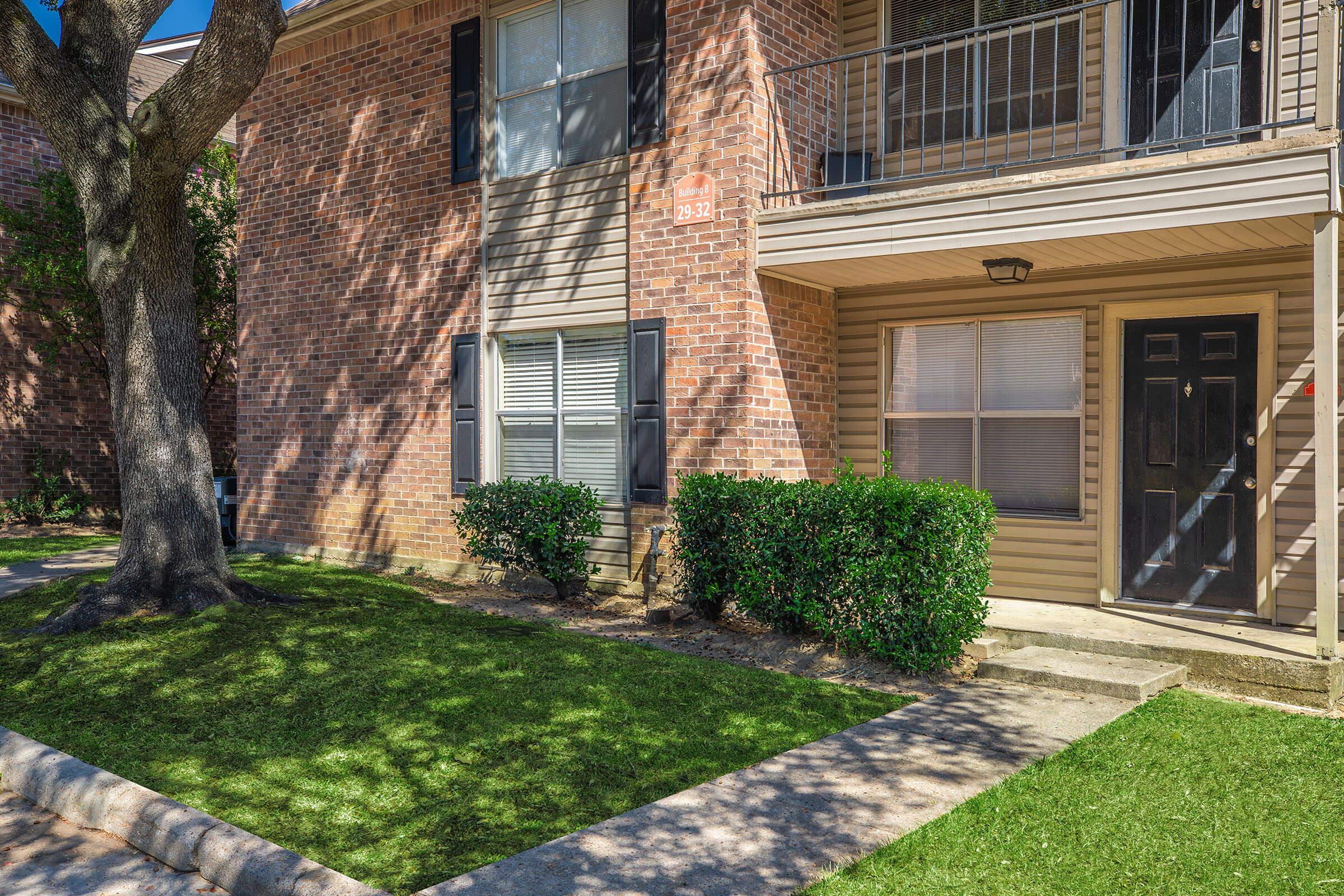 A corner view of a residential apartment building featuring a brick façade, black door, and front patio. There is a neatly trimmed hedge and a tree casting shadows on the lawn and walkway. Sunlight illuminates the scene, highlighting the well-maintained landscaping.