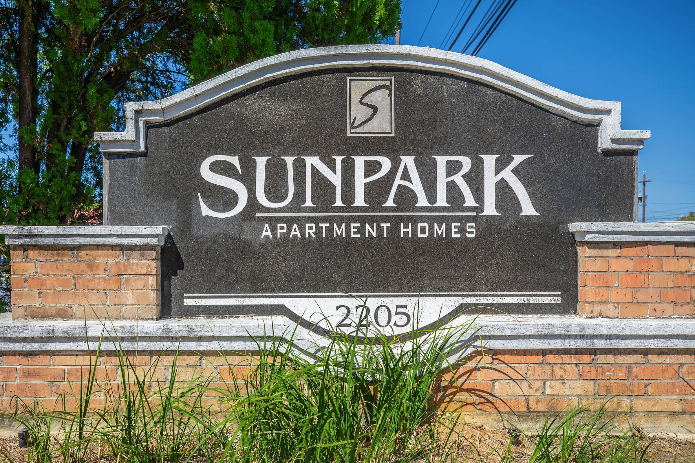 Sign for Sunpark Apartment Homes, featuring the name prominently in white text on a dark background. The address "2205" is displayed below. Surrounded by brick pillars and green grass, against a clear blue sky.