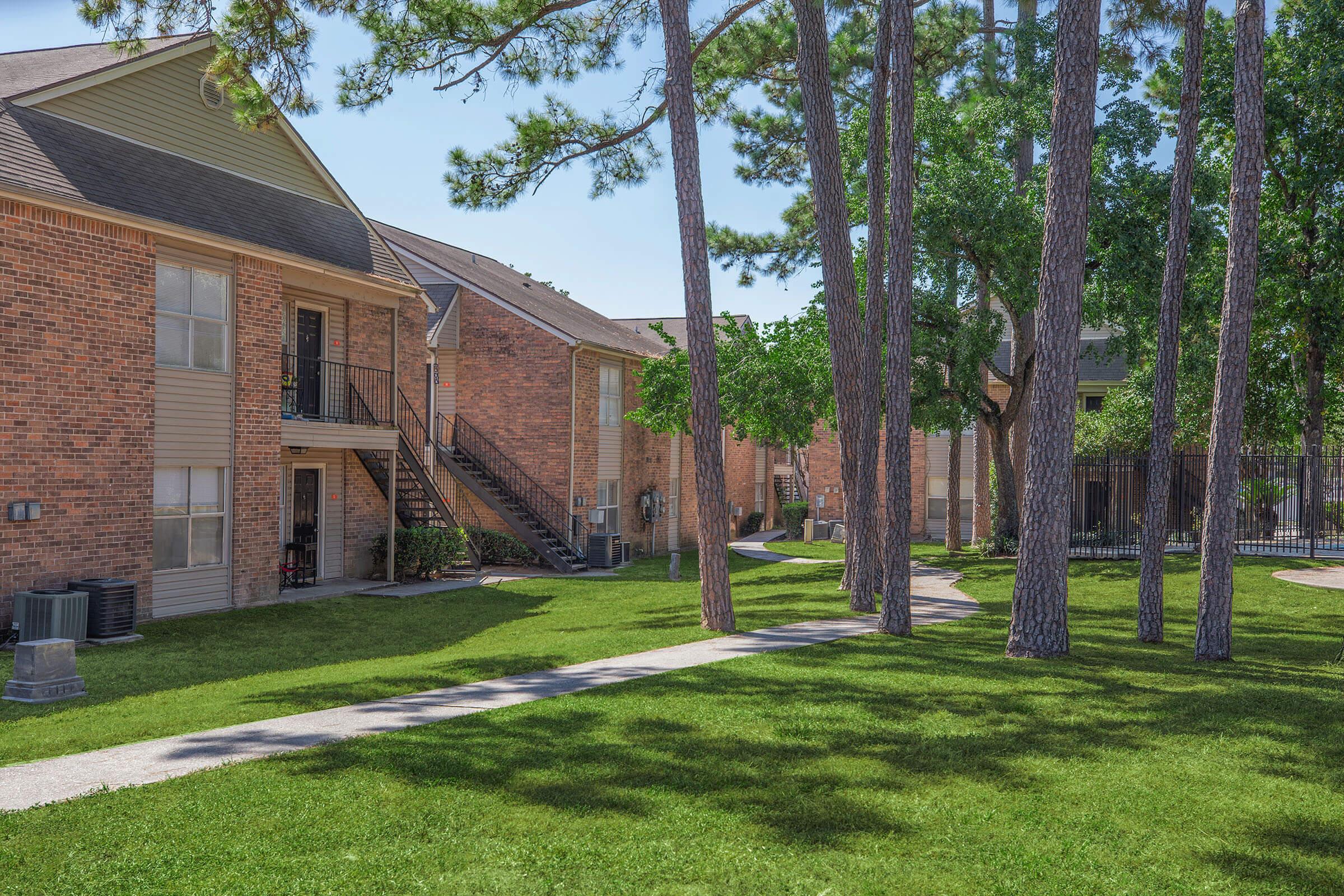 An outdoor view of a residential complex featuring two-story brick apartments. Green lawns are well-maintained, with a paved walkway winding through the area. Tall trees provide shade, and there is a glimpse of a fenced pool area in the background, indicating a pleasant living environment.