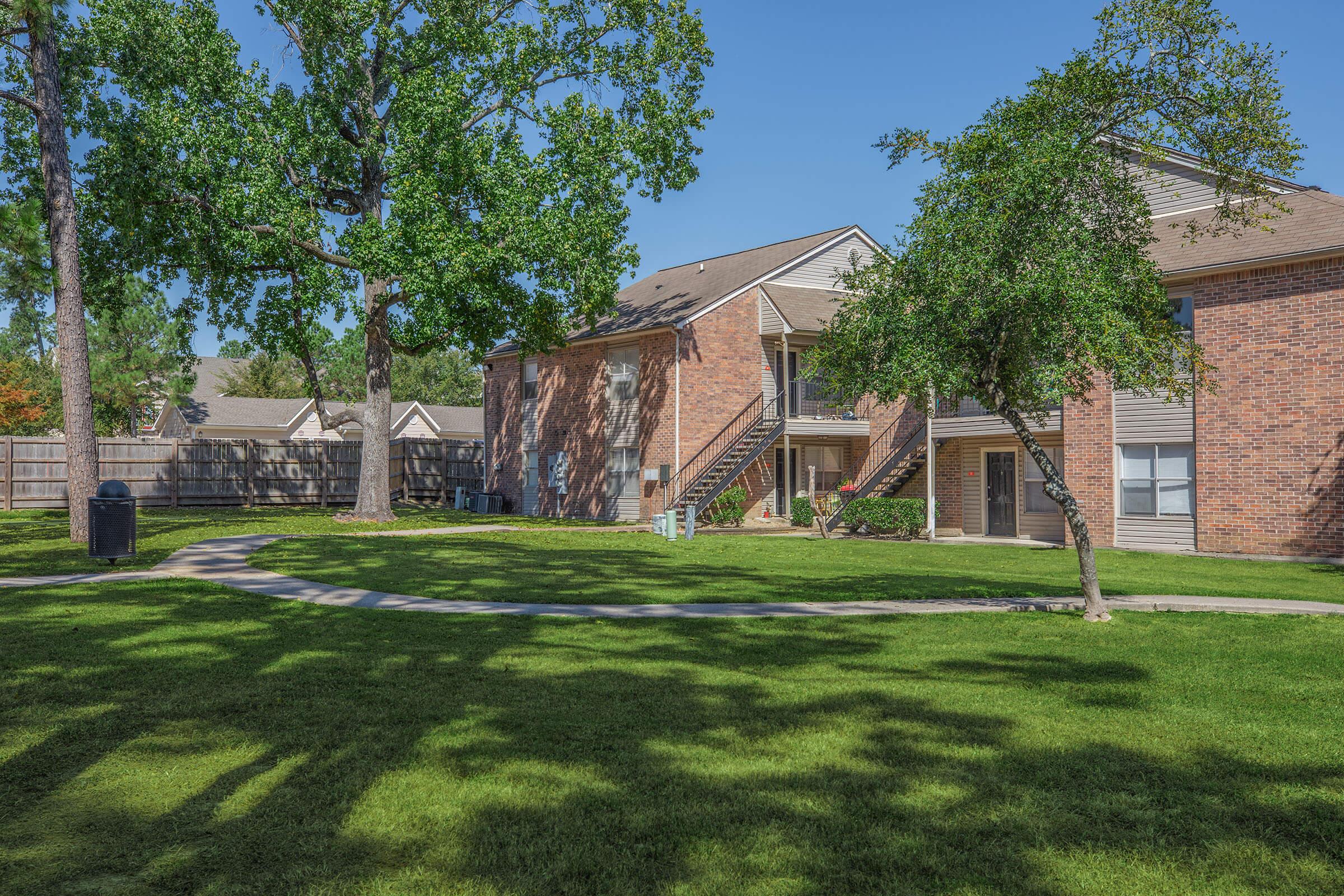 A well-maintained apartment complex featuring brick buildings surrounded by a grassy lawn and large trees. A pathway winds through the greenery, and there is outdoor seating and trash bins visible. The setting is bright and inviting under clear blue skies.