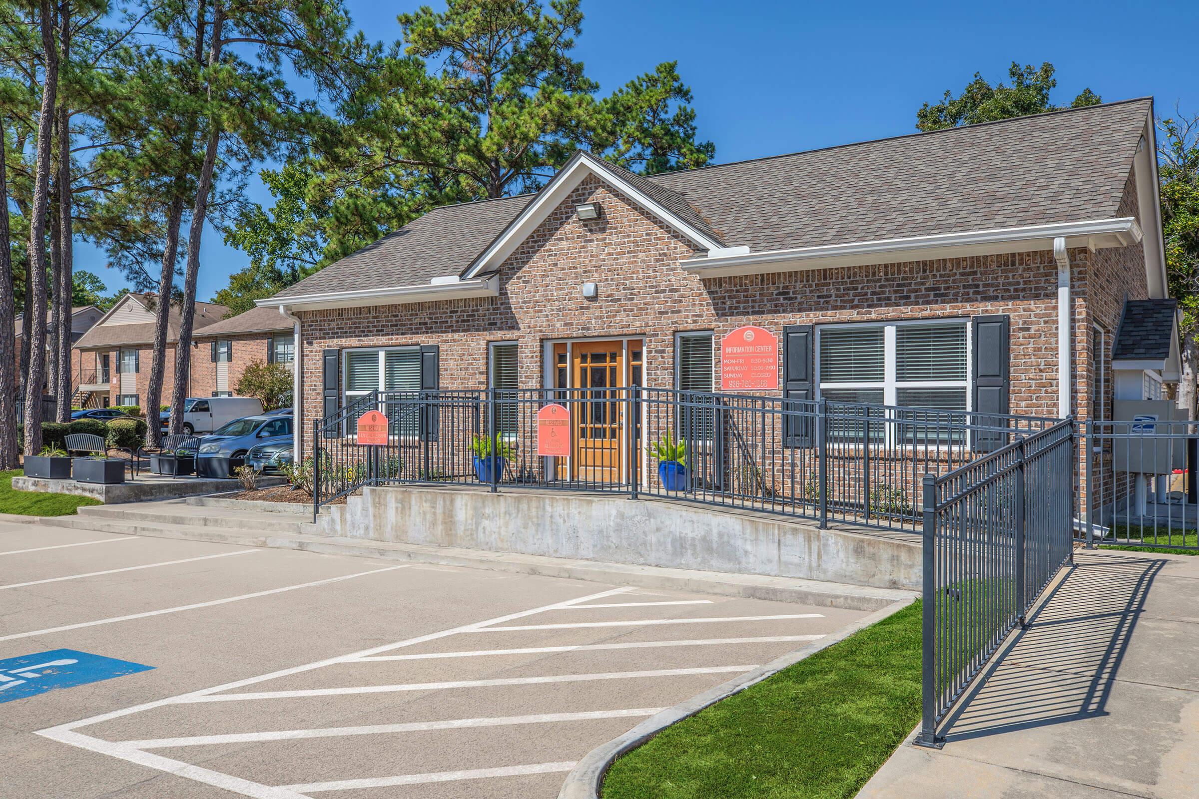 Brick building with large windows and a welcoming entrance. The facade features a mix of stone and wood elements, along with landscaped areas. A parking lot in front has marked spaces, and there are planters on either side of the entrance. Surrounding trees provide a natural backdrop.