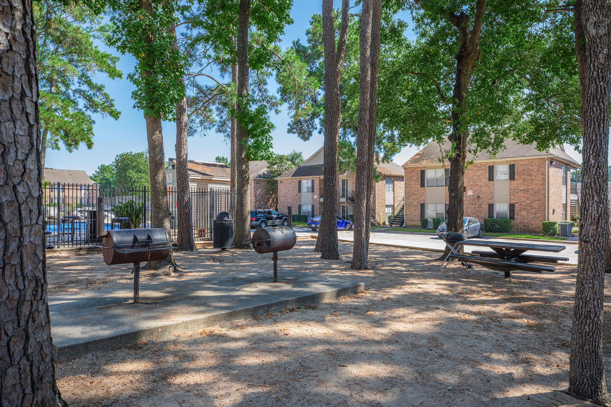 Shady outdoor area featuring several grills and a picnic table surrounded by tall trees. In the background, multiple brick apartment buildings are visible, along with a paved pathway and parking area. The scene is bright and inviting, ideal for outdoor gatherings.