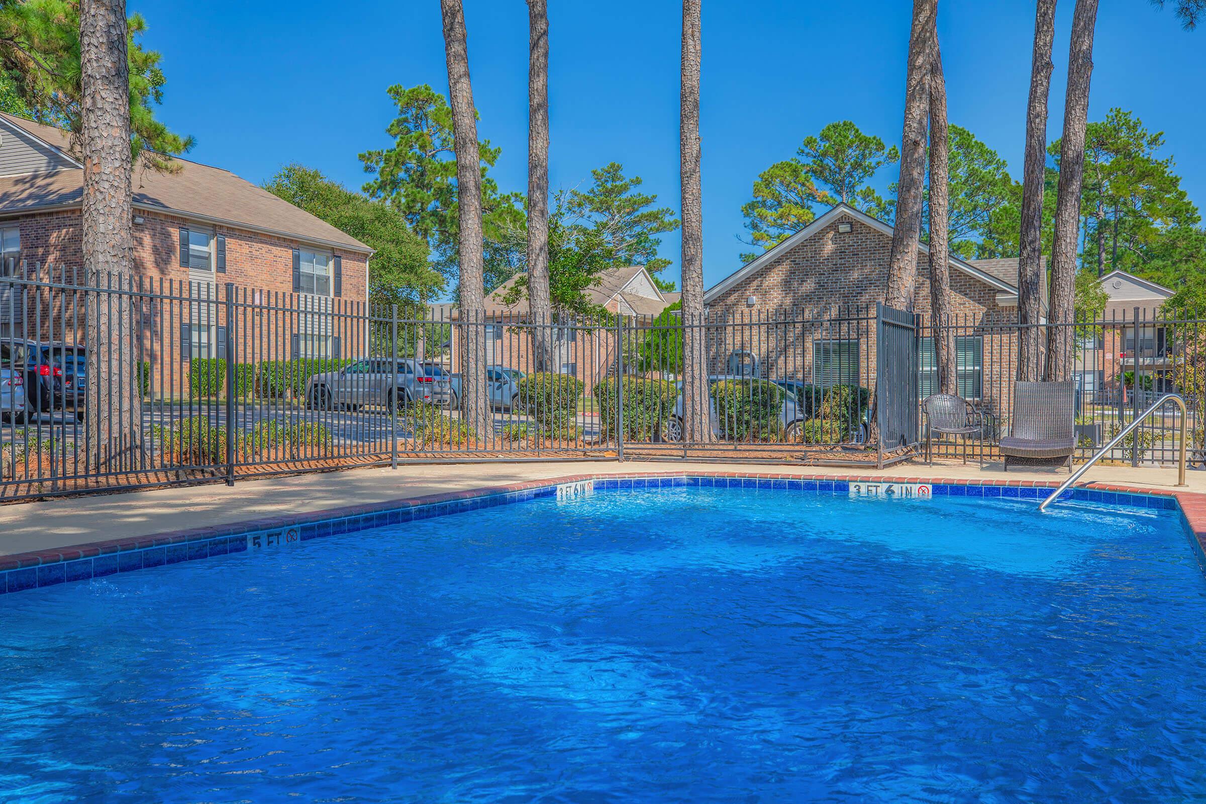 A clear blue swimming pool surrounded by tall trees and a wrought-iron fence. In the background, there are brick apartment buildings and parked cars. The sky is bright blue, indicating a sunny day. Comfortable lounge chairs are placed near the pool area.