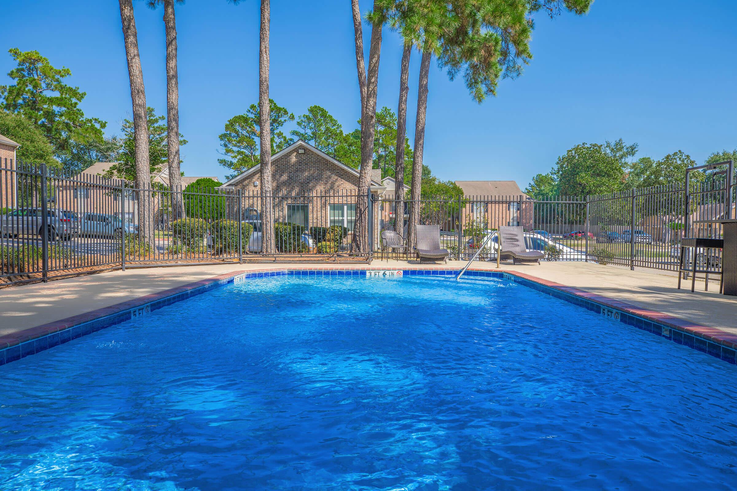 A clear blue swimming pool surrounded by a fenced area, with lounge chairs on the pool deck. In the background, residential buildings and tall trees can be seen under a bright blue sky. The scene conveys a relaxing outdoor environment.