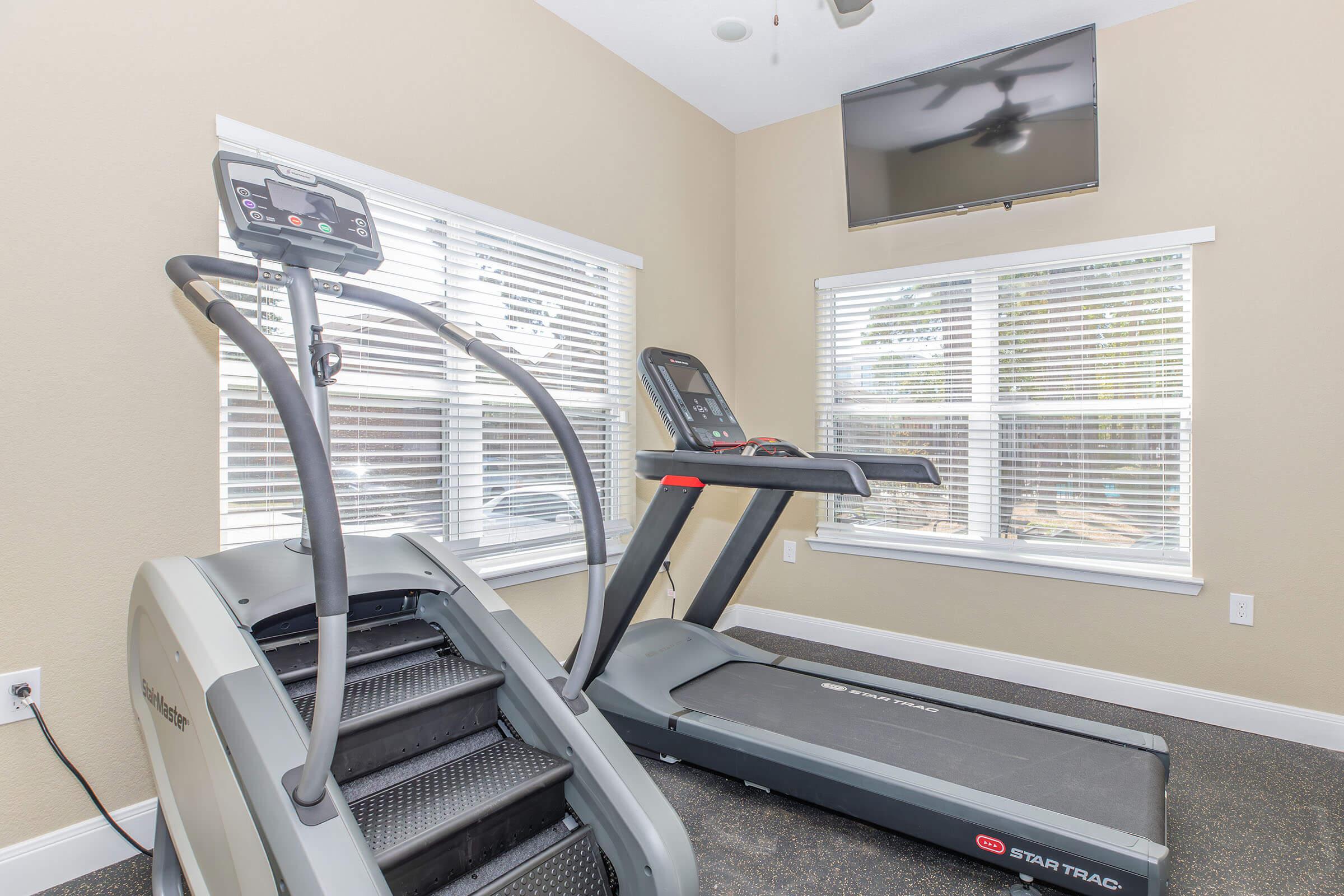 A fitness room featuring an elliptical machine and a treadmill, both facing a window with natural light. There's a wall-mounted television above the treadmill, and light-colored walls create a bright and inviting atmosphere.