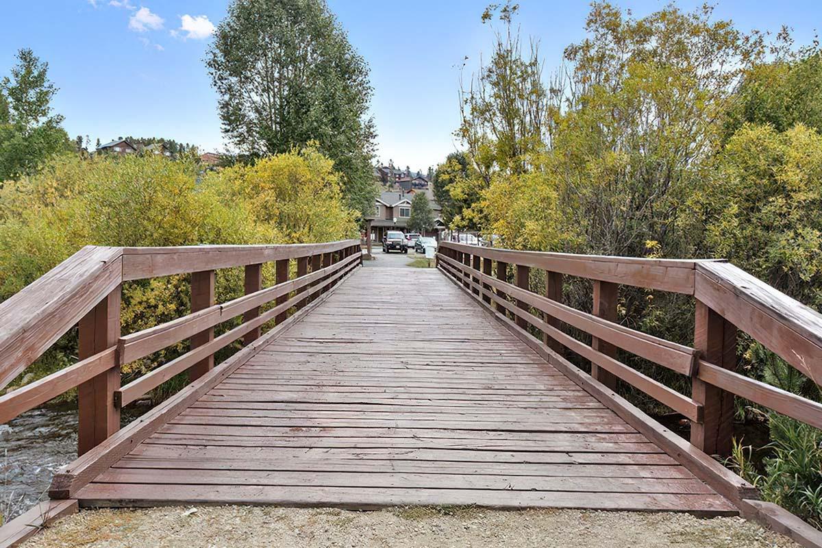 A wooden bridge leads into a scenic area with trees and a blue sky. The path on the bridge opens to a view of a parking area and buildings in the background, surrounded by autumn foliage. The peaceful landscape suggests a serene outdoor setting.