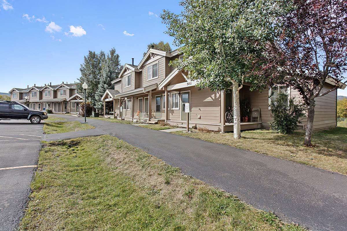 Row of residential houses with light-colored exteriors lining a paved walkway. Green lawns and trees are visible in the foreground, and a parking area is adjacent to the houses. The sky is clear, indicating a sunny day.