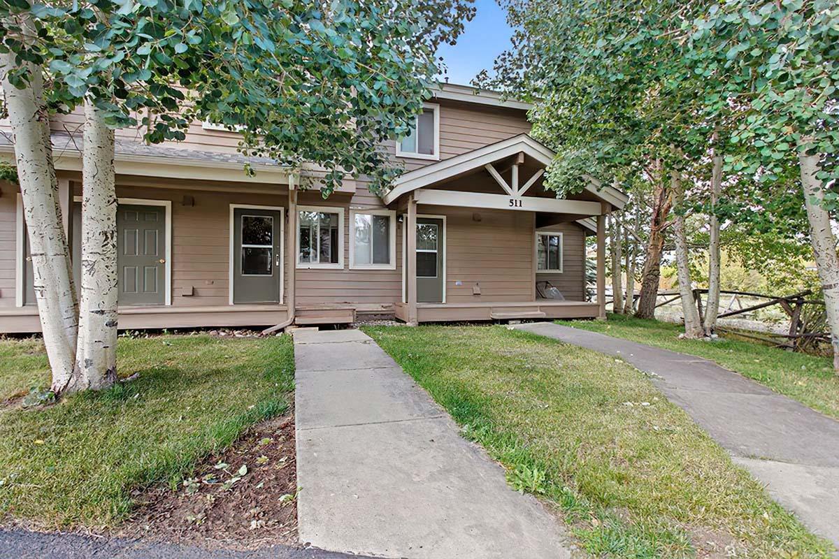 Two-story wooden house with a light brown exterior, featuring a covered porch and large windows. The front yard has green grass and trees. A concrete path leads to the entrance, with a clear blue sky in the background. The house number 511 is displayed above the door.