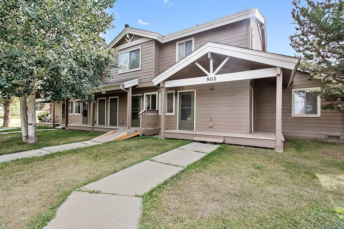 Exterior view of a two-story townhouse with brown siding, featuring a sloped roof, a wide porch entrance, and a ramp for accessibility. The grassy area in front has concrete walkways leading to the doors, surrounded by trees and shrubs against a clear blue sky.