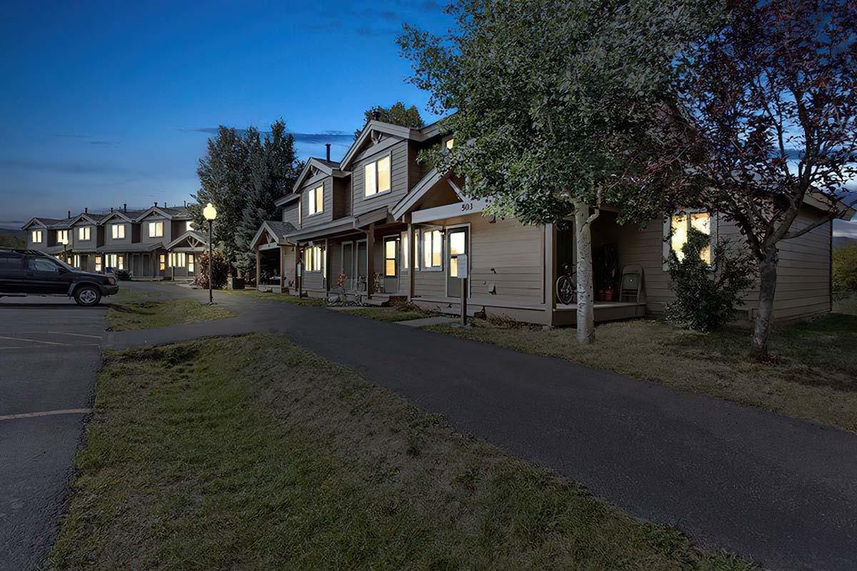A row of residential houses at dusk, with soft lights illuminating the windows. Trees are lined along the pathway, and a well-maintained lawn is visible in front of the homes. The scene conveys a peaceful suburban atmosphere in the evening.