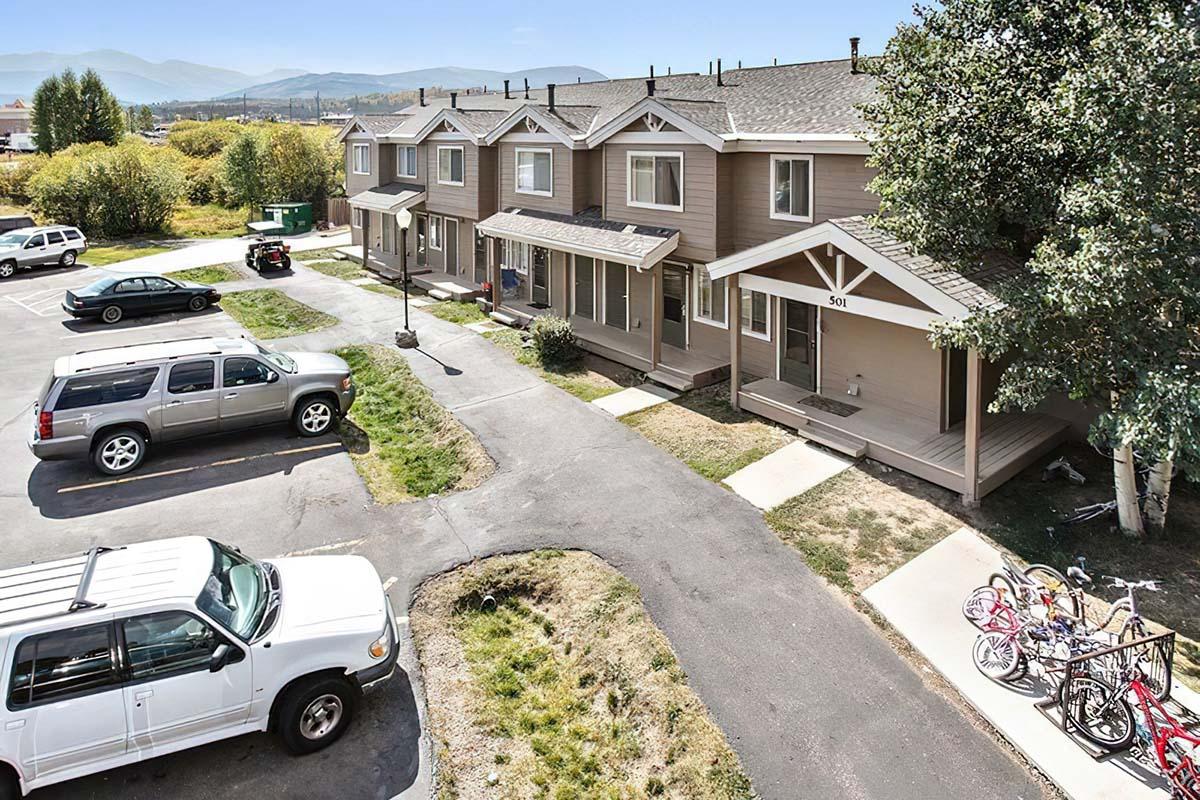 A view of a residential area featuring several townhouse-style units with porches. There are parked cars in the foreground, along with bicycles near the entrance of the buildings. The surroundings include grassy areas and trees, set against a backdrop of mountains and a clear blue sky.