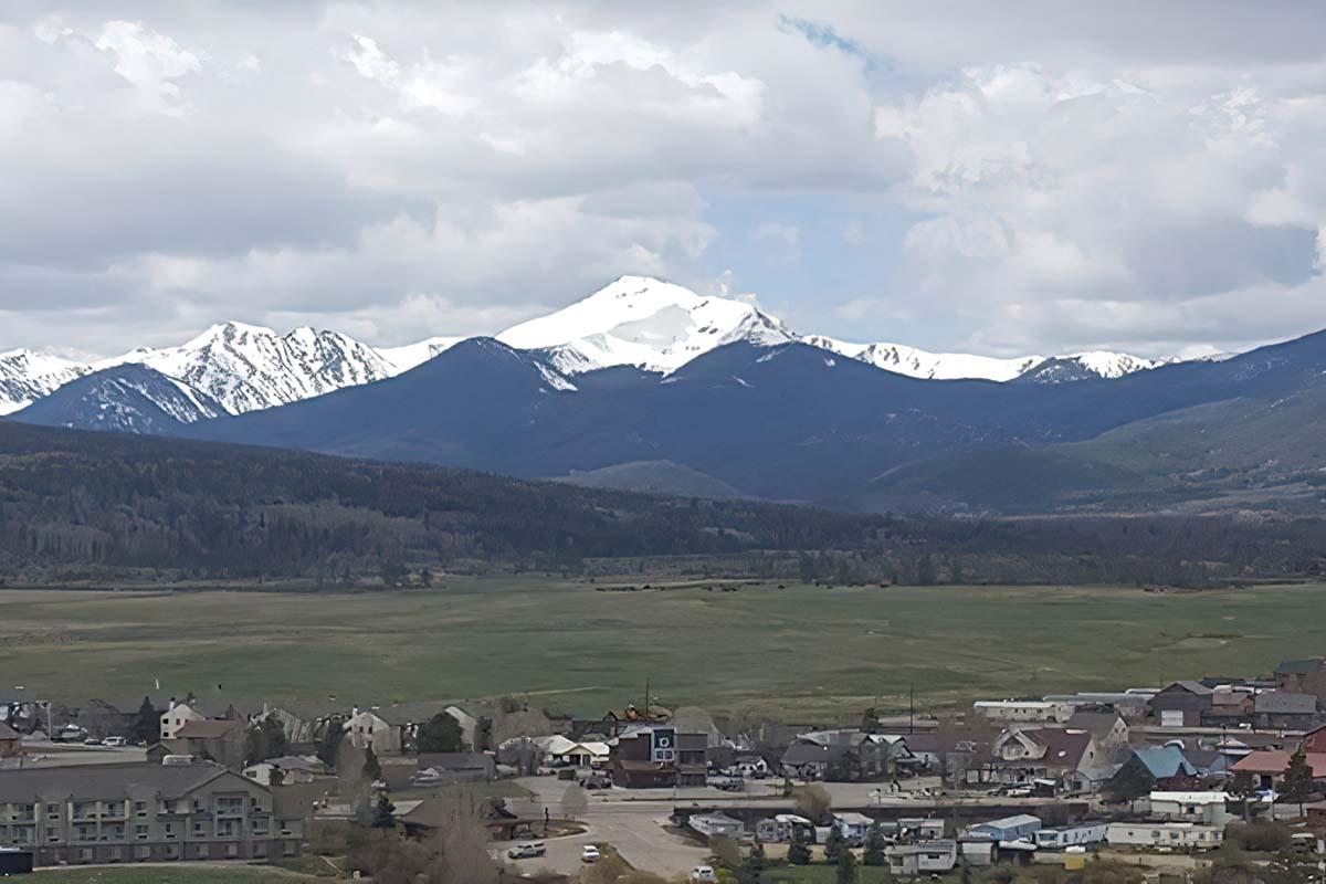 A panoramic view of a mountainous landscape featuring snow-capped peaks under a partly cloudy sky. In the foreground, a small town with multiple buildings is visible, surrounded by grassy fields and forested areas. The mountains in the background create a dramatic backdrop.