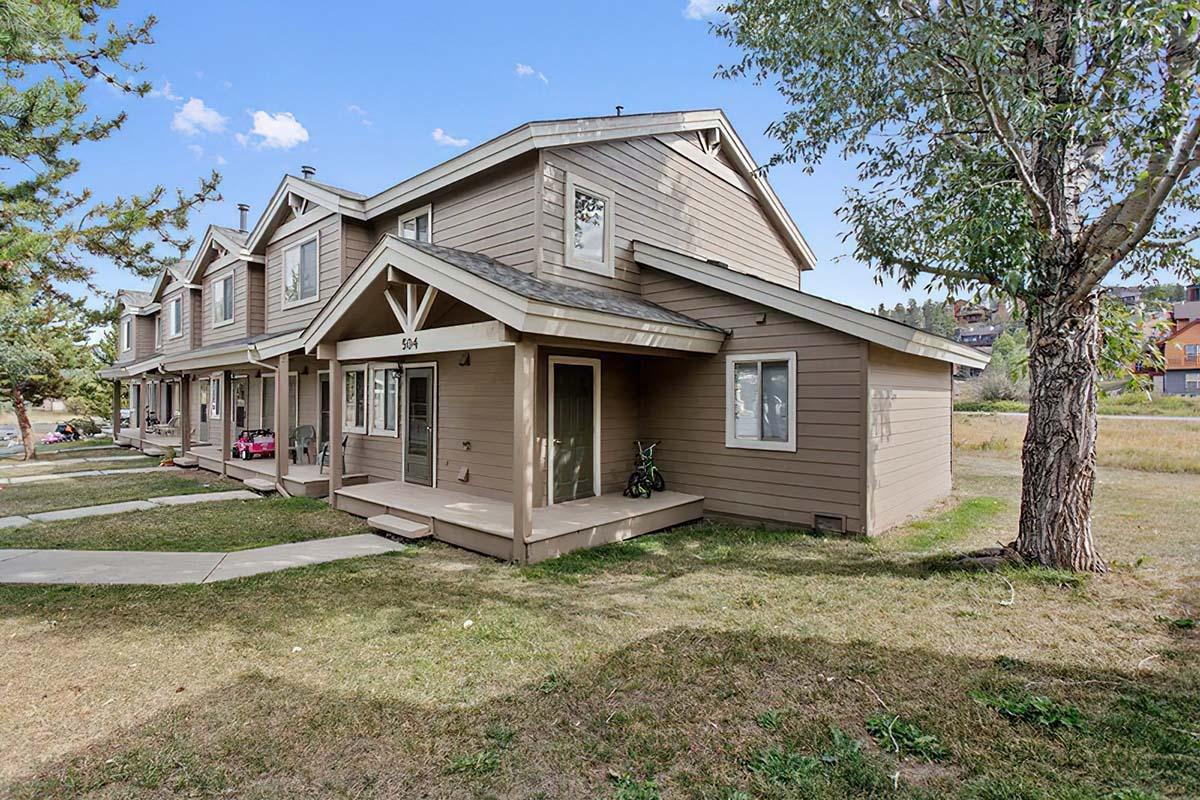 A row of two-story townhouses with a wooden exterior, featuring porches and front yards. The image shows pathways leading to the entrance, surrounded by grassy areas and a few trees. Ideally located in a residential neighborhood with a clear blue sky in the background.